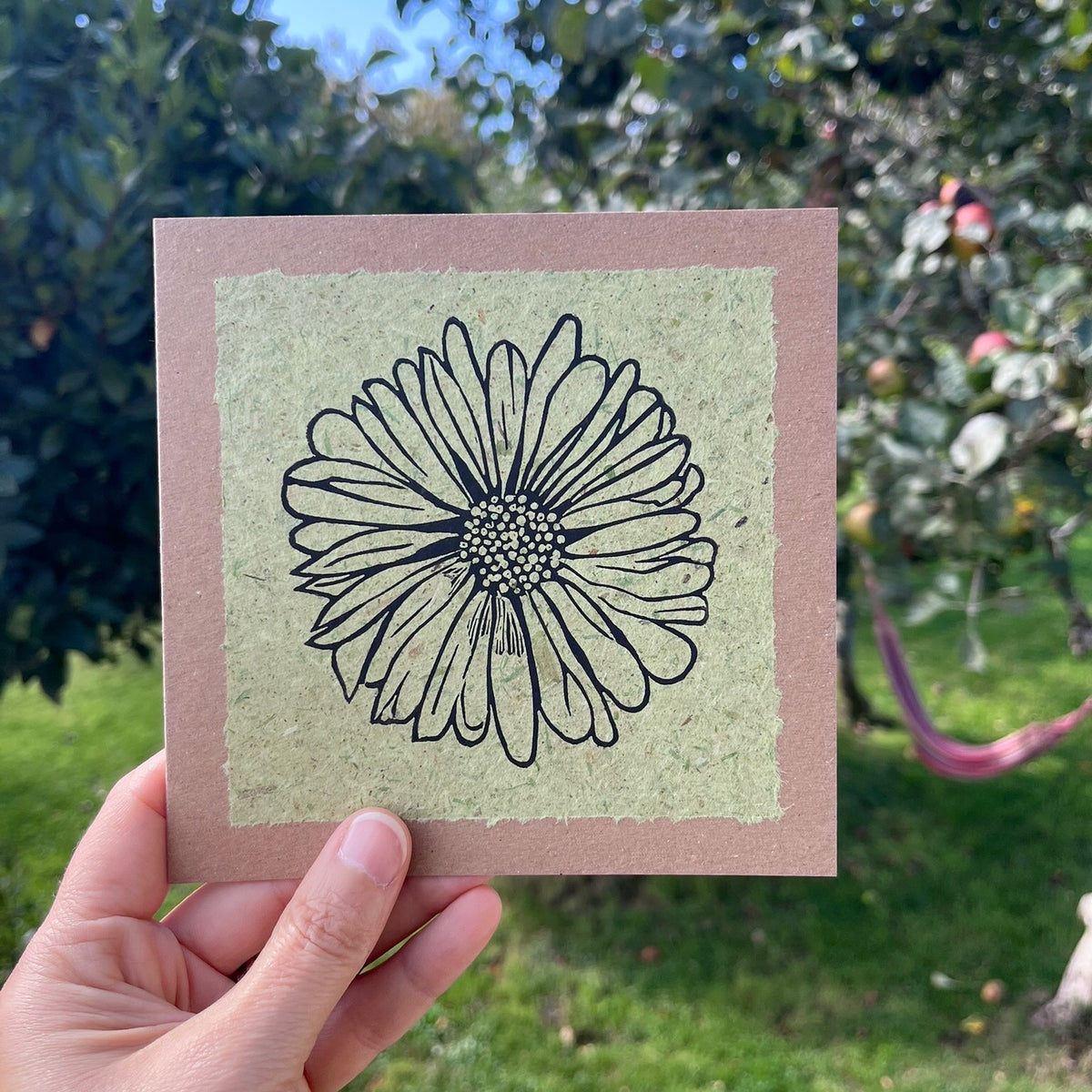 A hand holding a square handmade art card featuring a black ink lino print of a daisy flower on a textured light green handmade paper, mounted on a brown kraft greetings card, held against a garden backdrop.