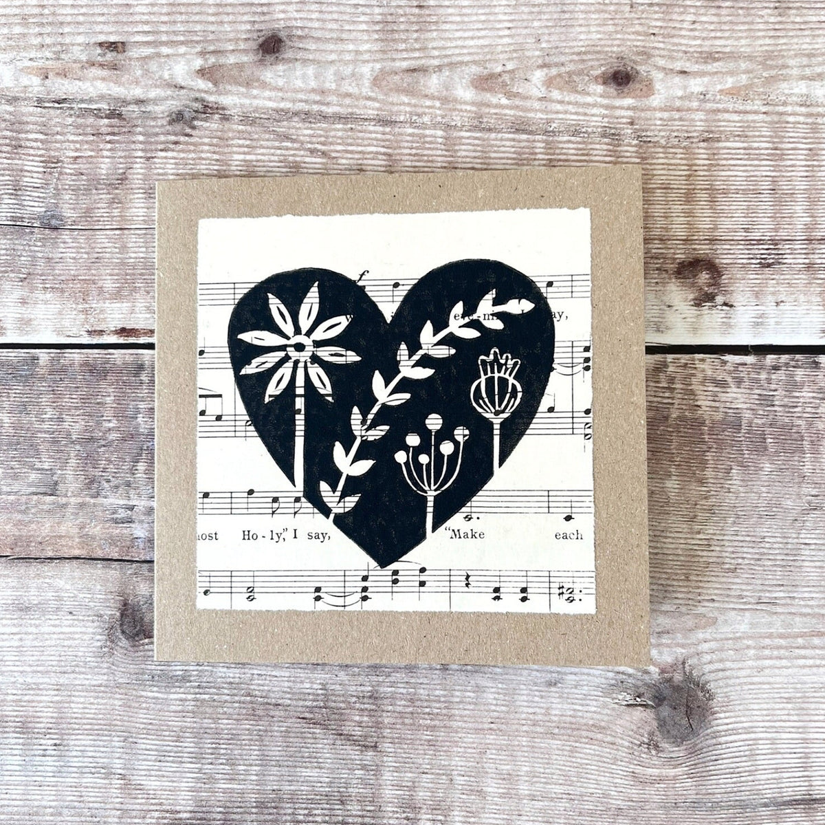 Square kraft greeting card with a black floral heart lino print on vintage music paper mounted on the front. Laid on a wooden background.