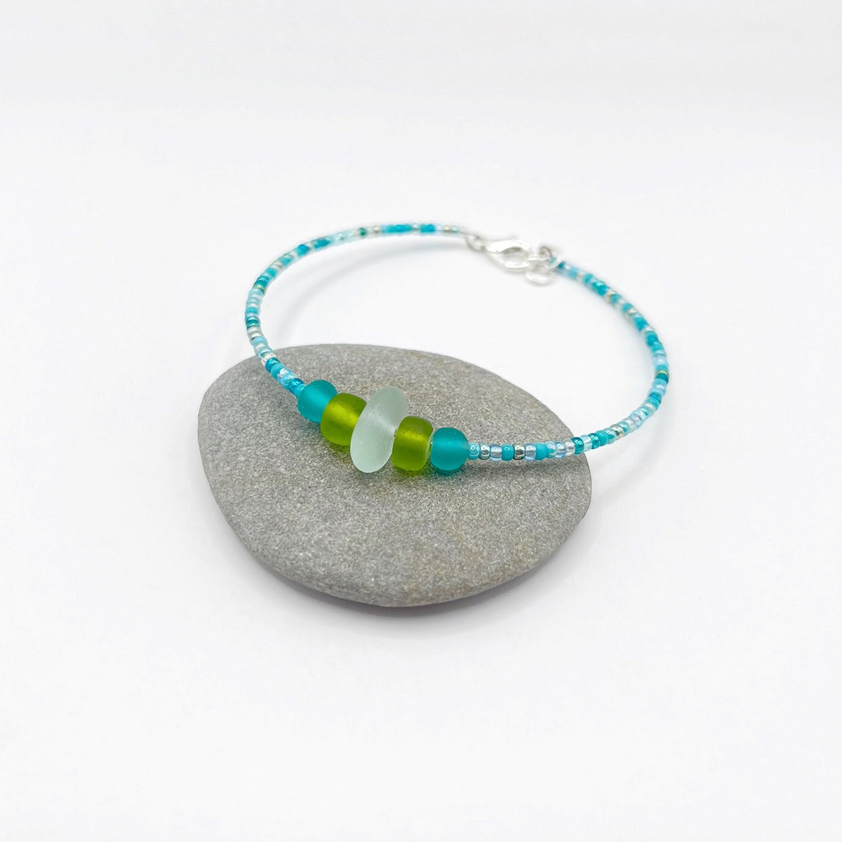 A turquoise beaded bracelet with lime and sea green frosted glass beads and a piece of sea foam coloured Cornish sea glass, finished with a silver plated lobster clasp and laid on a grey pebble.
