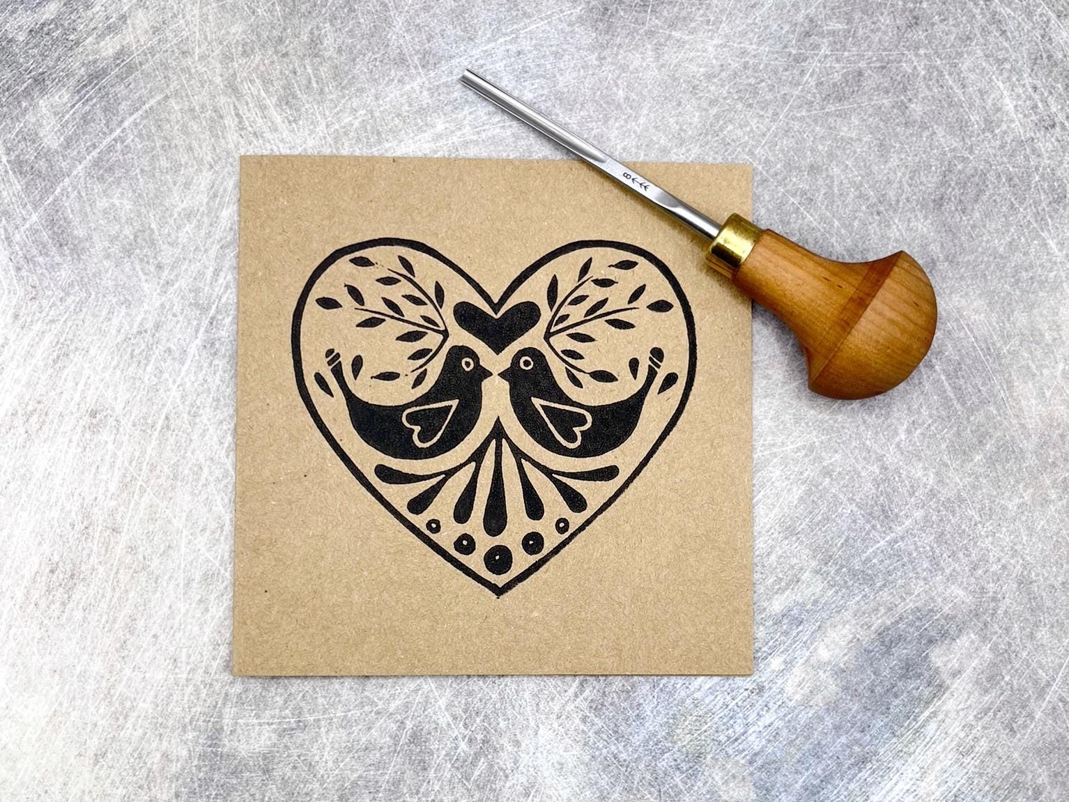 Kraft card with an original lino print heart featuring two birds. Photo includes lino cutting tool used laid on a textured background.