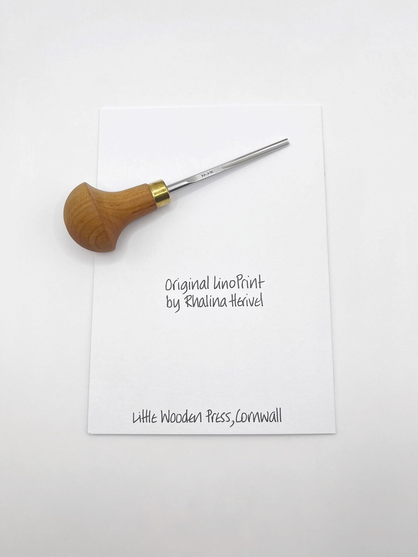 Showing the back of the white card, featuring text 'Original linocut by Rhalina Herivel' and 'Little Wooden Press, Cornwall'. Photo includes lino cutting tool used.