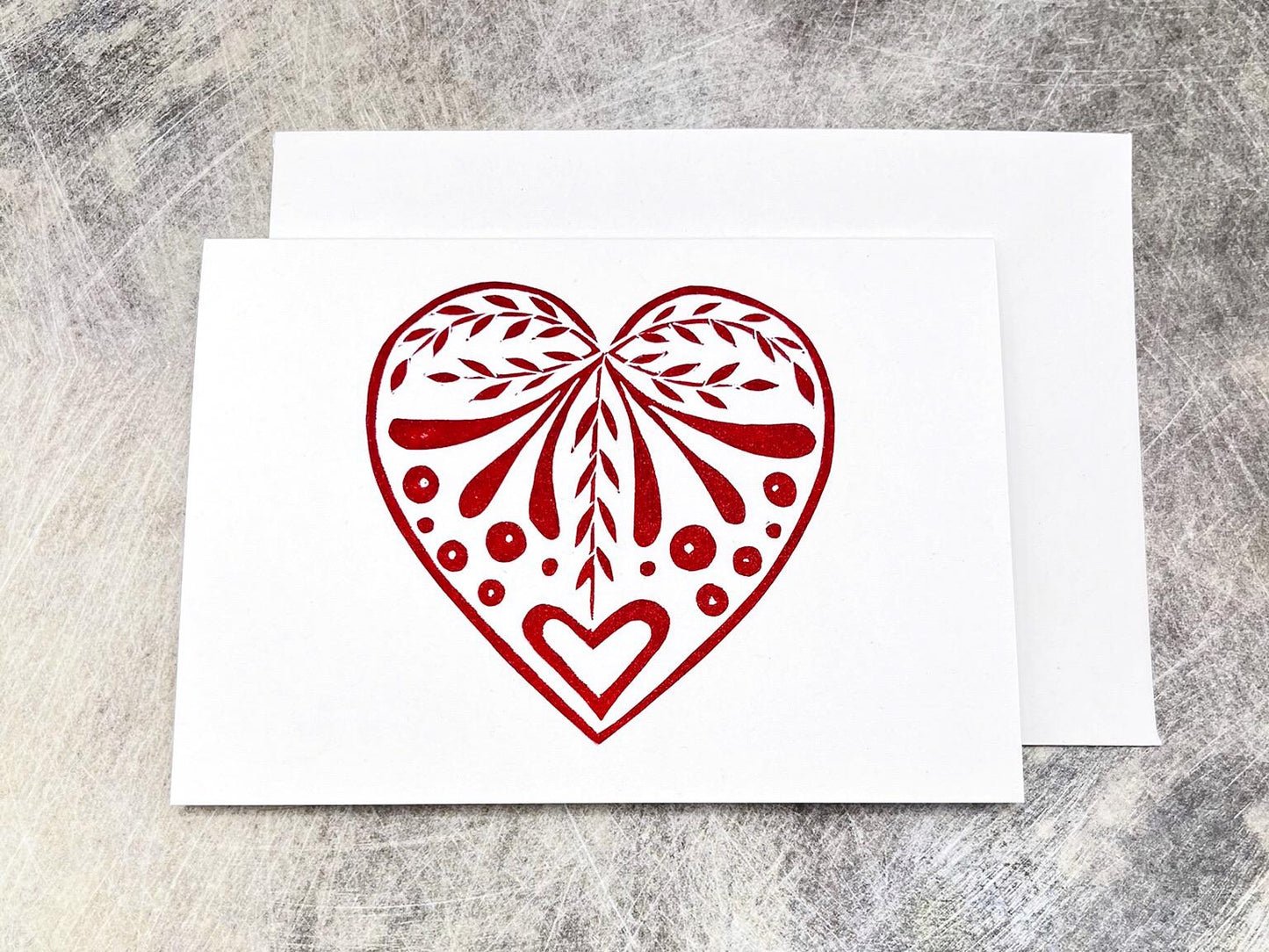 White card with an original lino printed red heart design laid on a textured surface. 