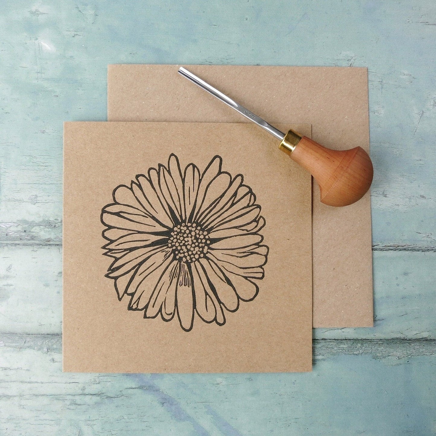 Kraft paper card with an original daisy lino print laid on a textured surface. Photo includes lino cutting tool