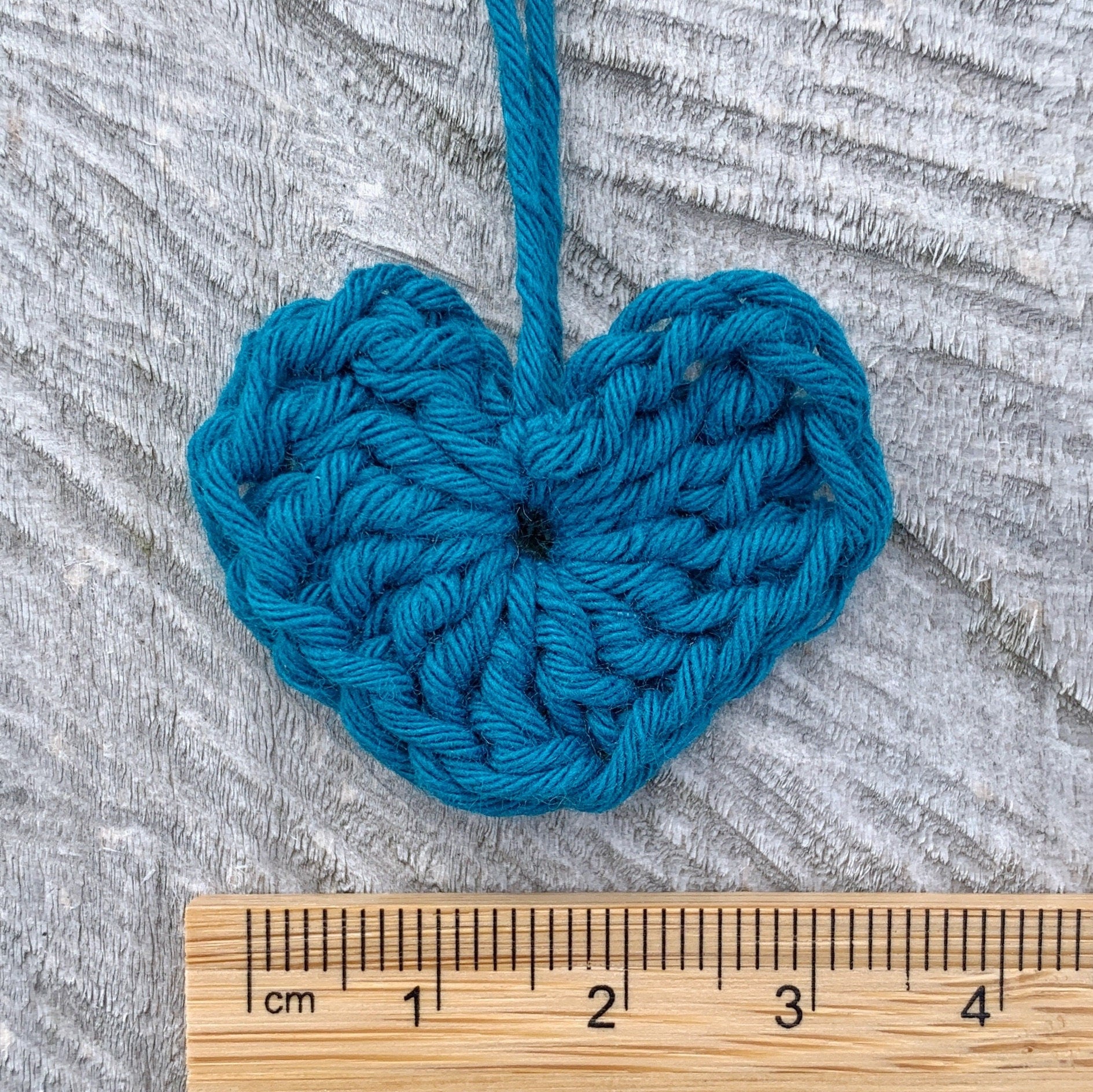 Teal crocheted heart with a wooden ruler for scale on a textured grey background