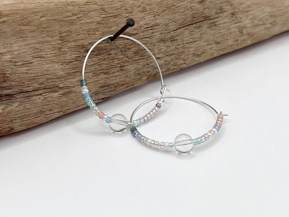 Pair of pastel seed bead and clear crystal hoop earrings hanging on driftwood against a white surface