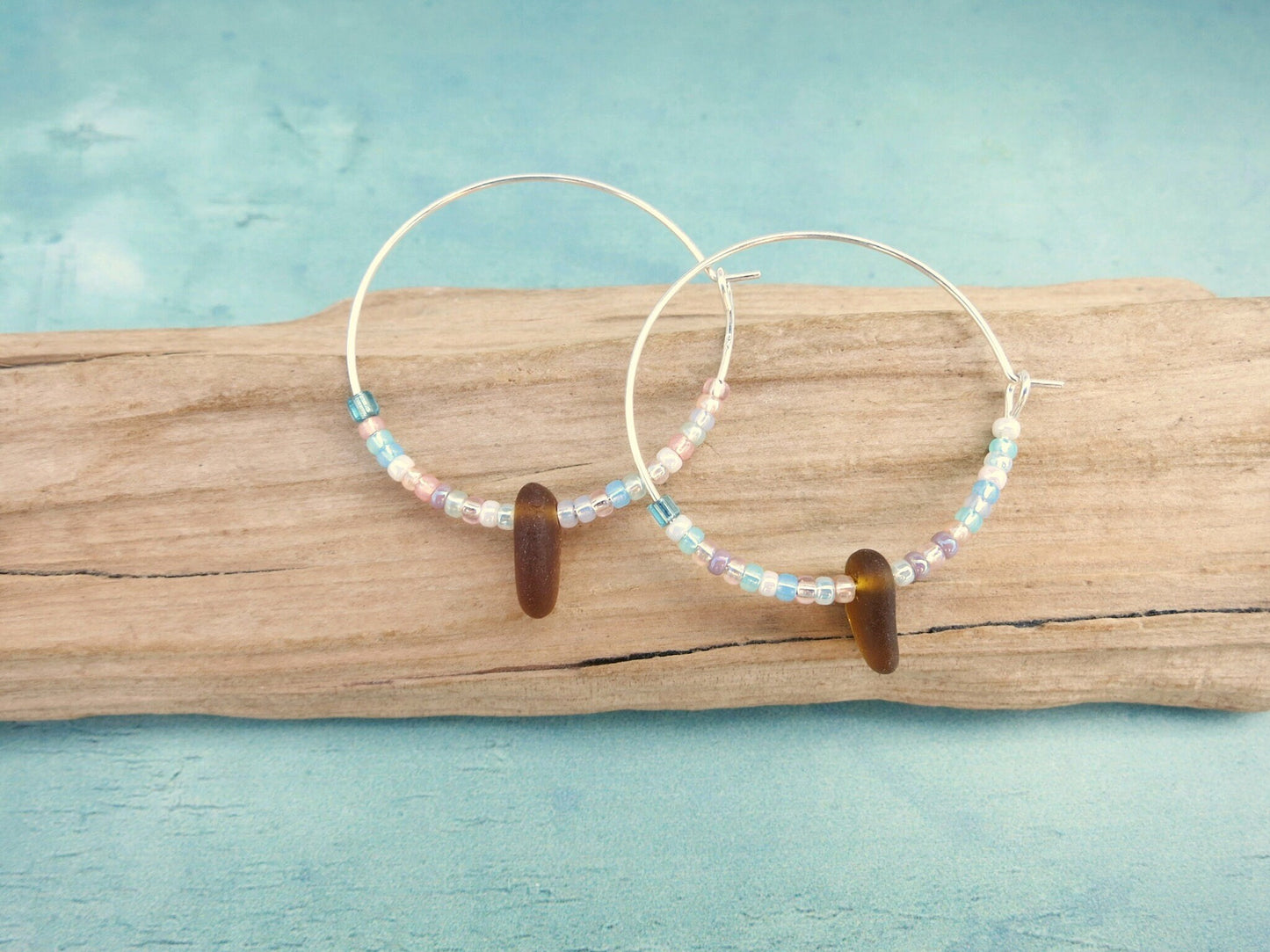 Silver hoop earrings with pastel seed beads and burnt orange sea glass laid on a piece of driftwood against a light blue background