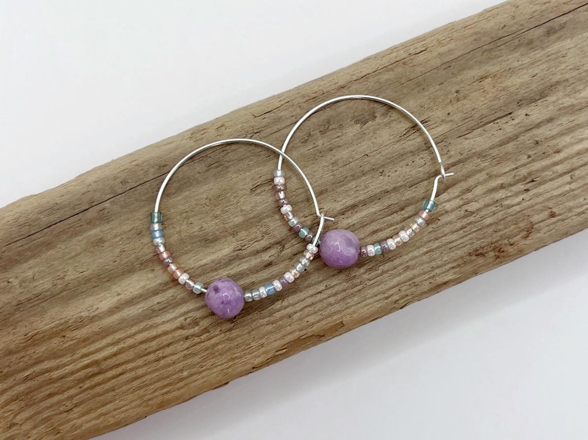 Pair of silver hoop earrings with lilac Jasper and seed beads laid on driftwood against a light surface.