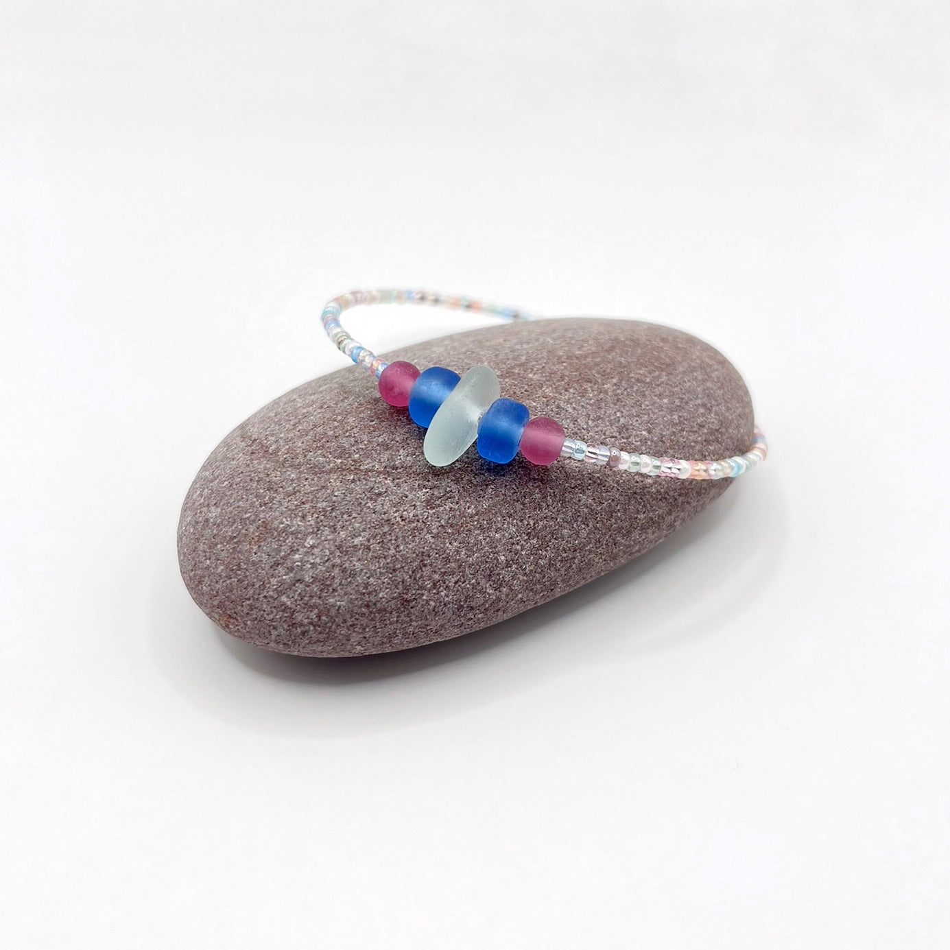 A pastel beaded bracelet with pink and blue frosted glass beads and a piece of sea foam coloured Cornish sea glass, finished with a silver plated lobster clasp and laid on a purple pebble.