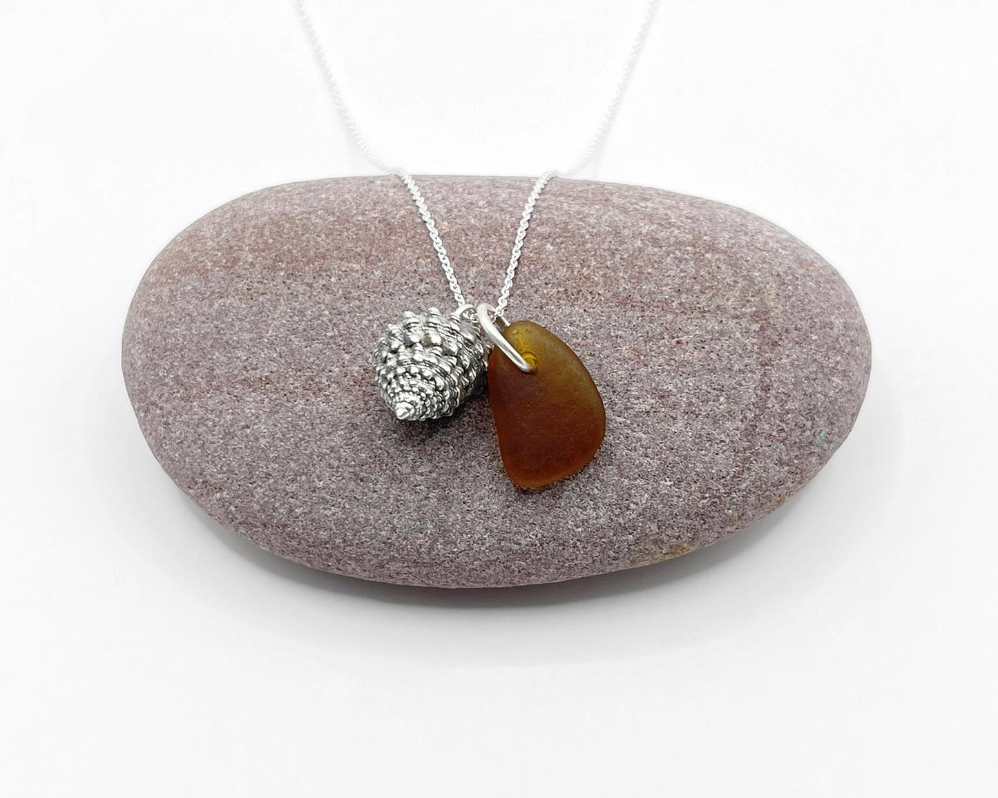 Necklace with burnt orange sea glass and silver shell pendant laid on a purple  pebble against a white background