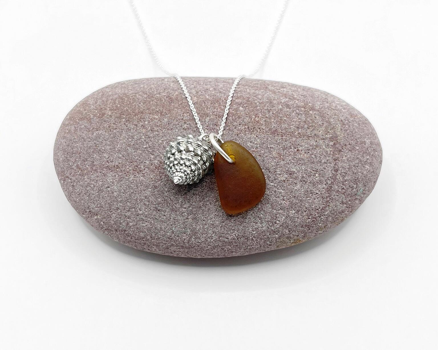 Necklace with burnt orange sea glass and silver shell pendant laid on a purple  pebble against a white background