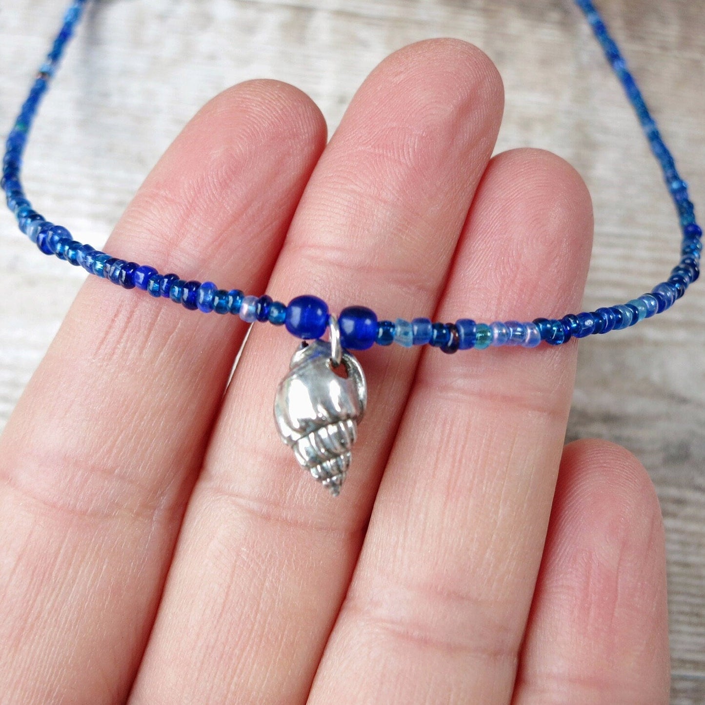 Blue seed bead necklace with a silver shell charm held on a hand against a neutral background