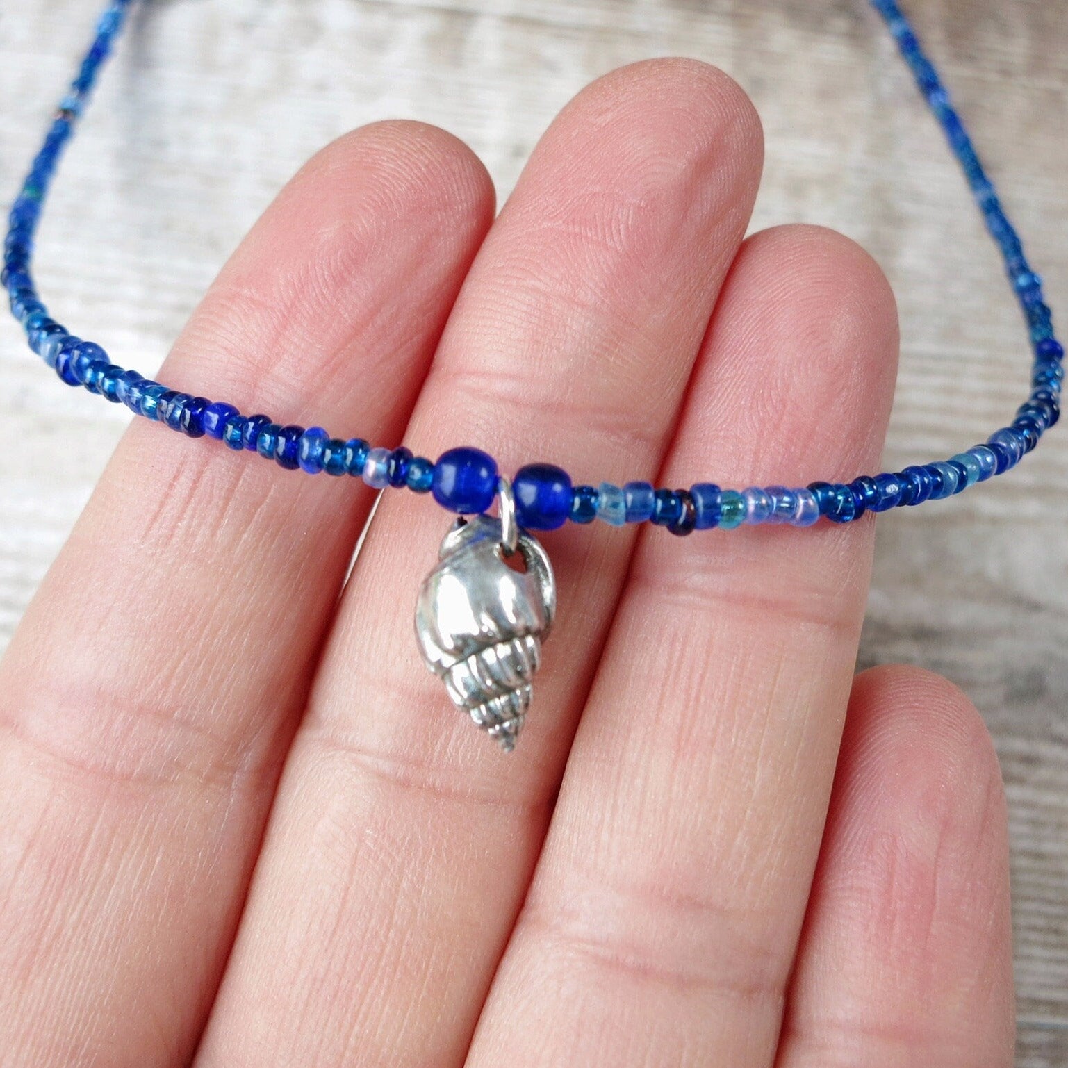 Blue seed bead necklace with a silver shell charm held on a hand against a neutral background