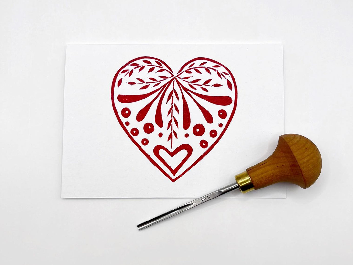 White card with an original lino printed red heart design stood on a white surface. Photo includes lino cutting tool.