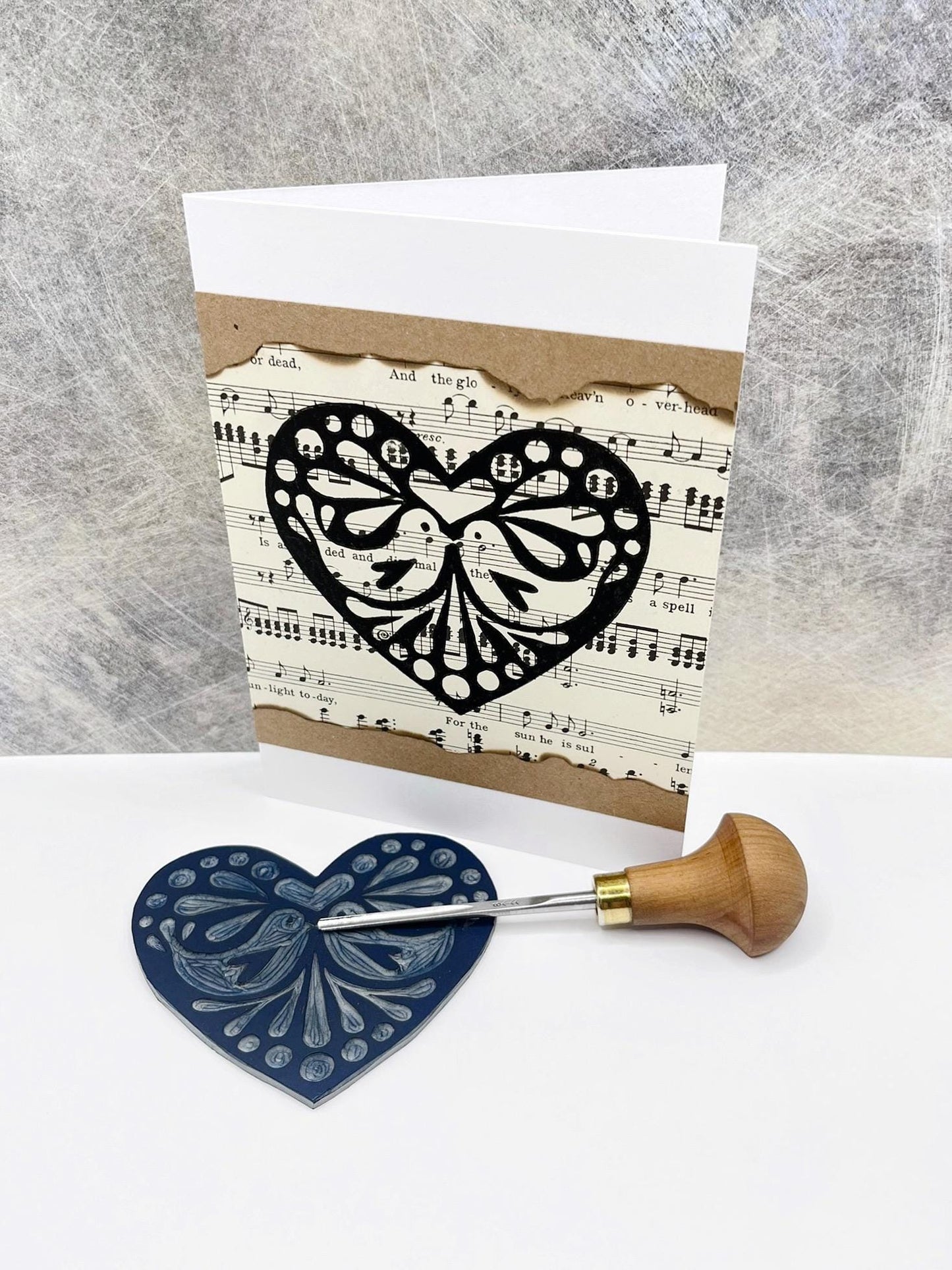 Card with original lino printed black heart design music sheet, stood against a textured background. Photo includes lino and cutting tool.