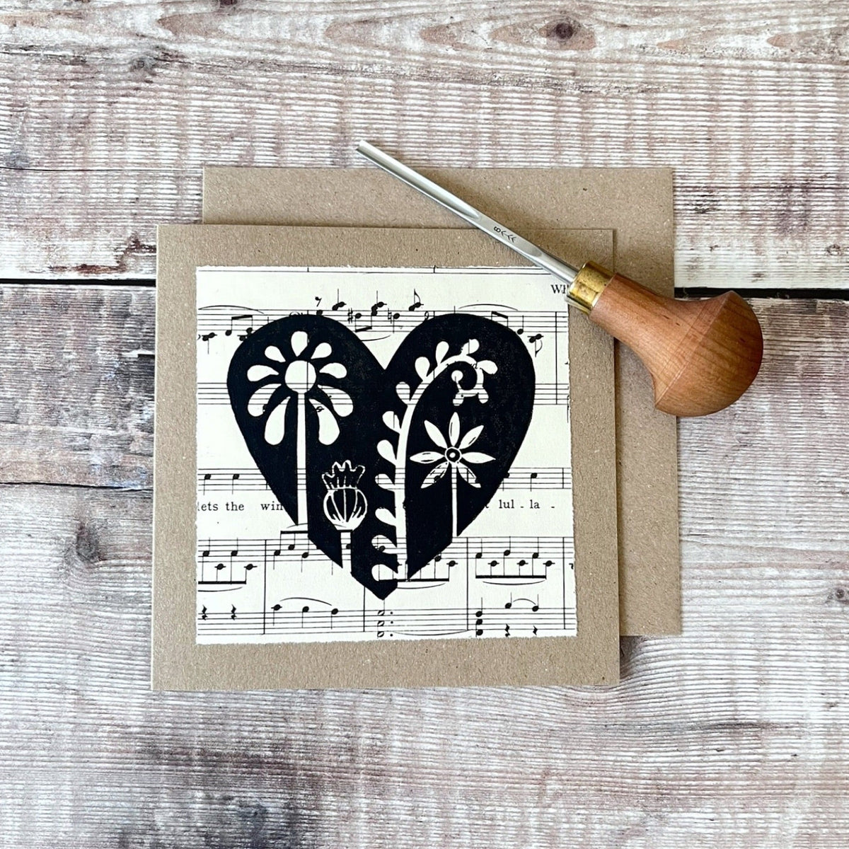 Square kraft greeting card with a black floral heart lino print on vintage music paper mounted on the front. Laid on a matching envelope on a wooden background. Photo includes the lino cutting tool used.
