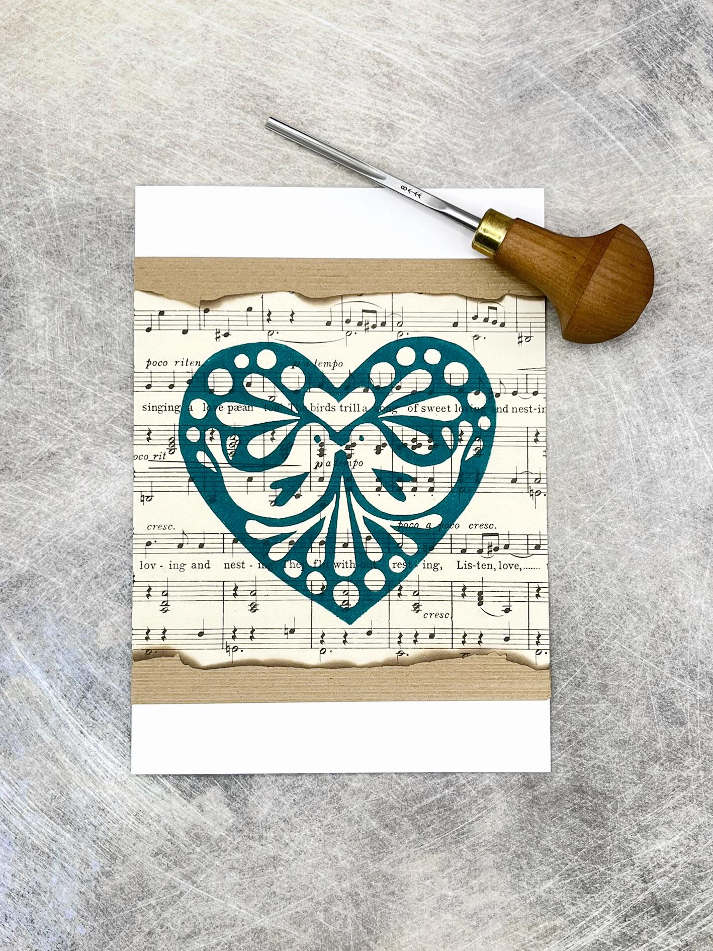 Card with an original linomprinted heart design on sheet music. Included in the photo is the lino cutting tool used laid on a textured background.