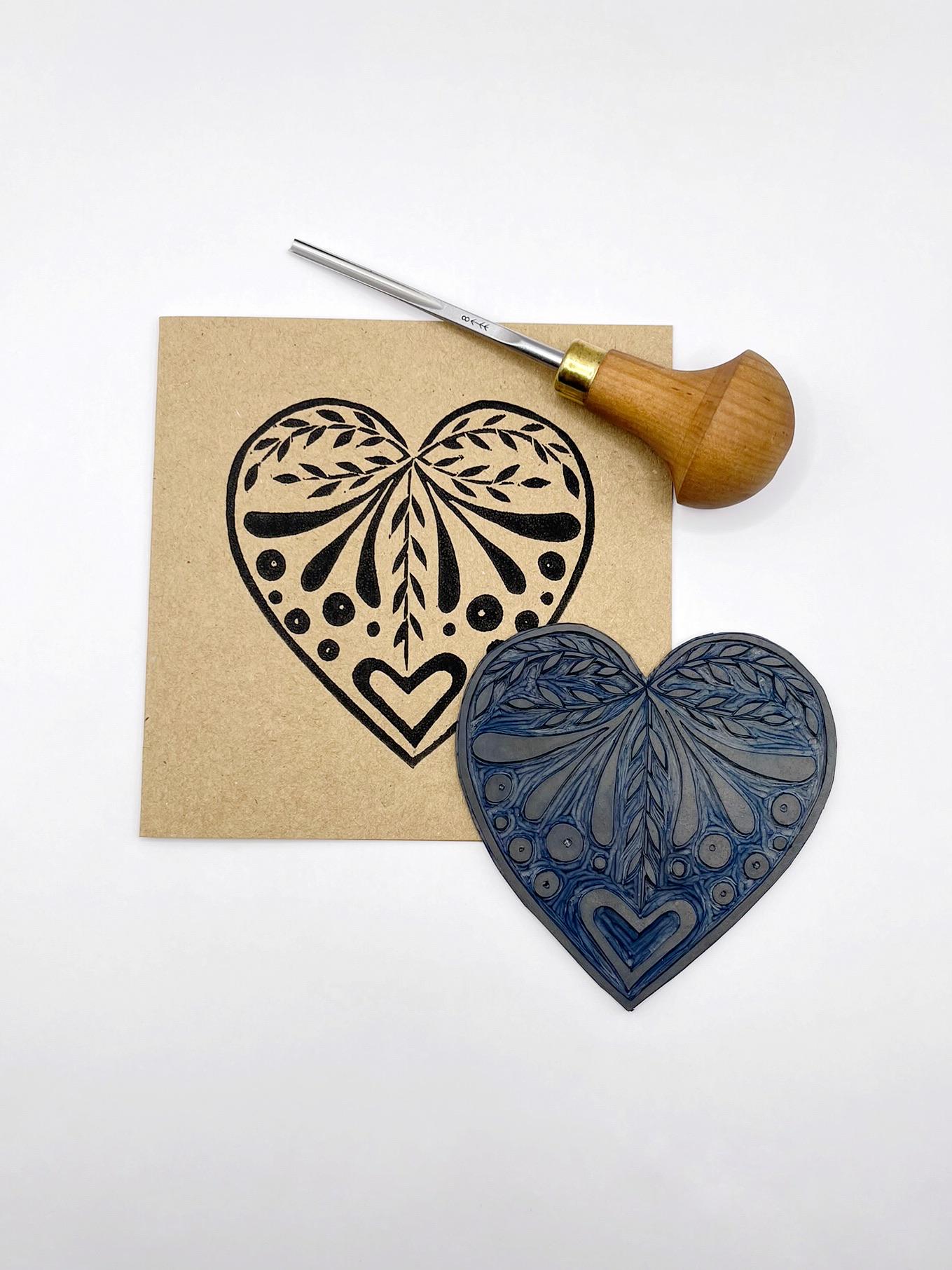 Card with an original lino printed black heart design on Kraft card laid on a white surface. Photo includes lino and cutting tool used.