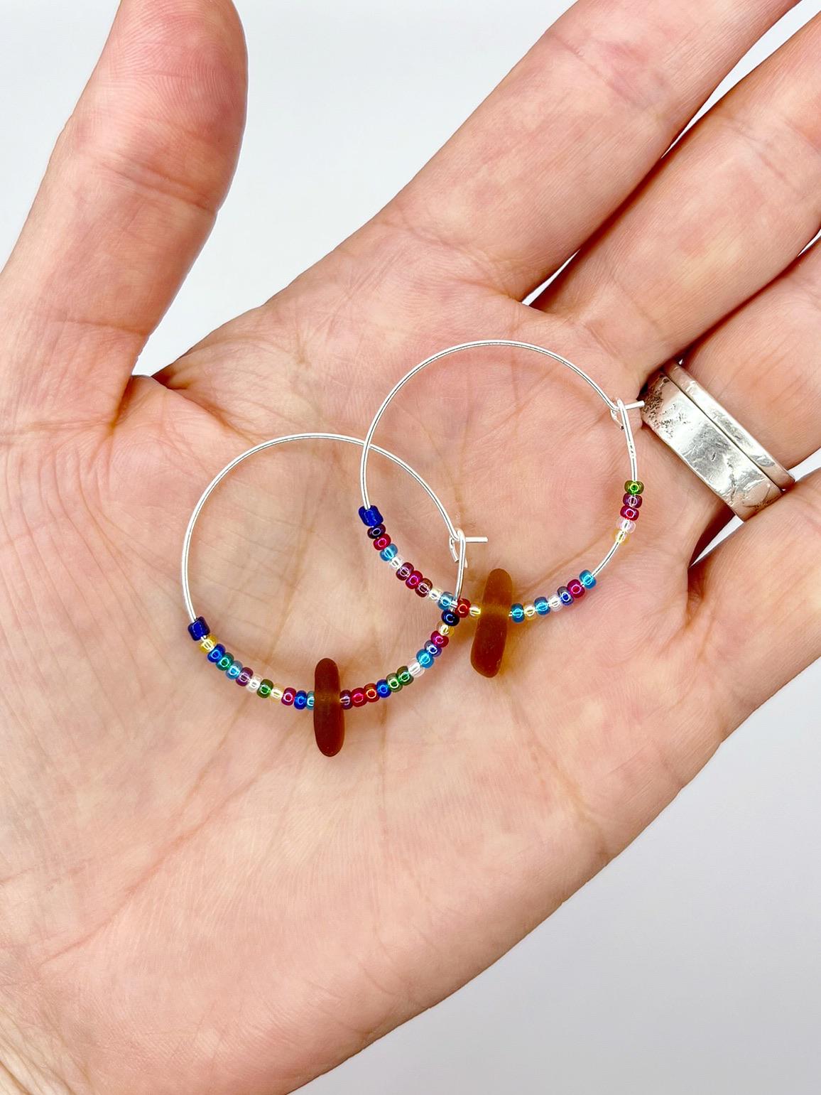 Rainbow seed bead and burnt orange sea glass hoop earrings held in a hand against a white background