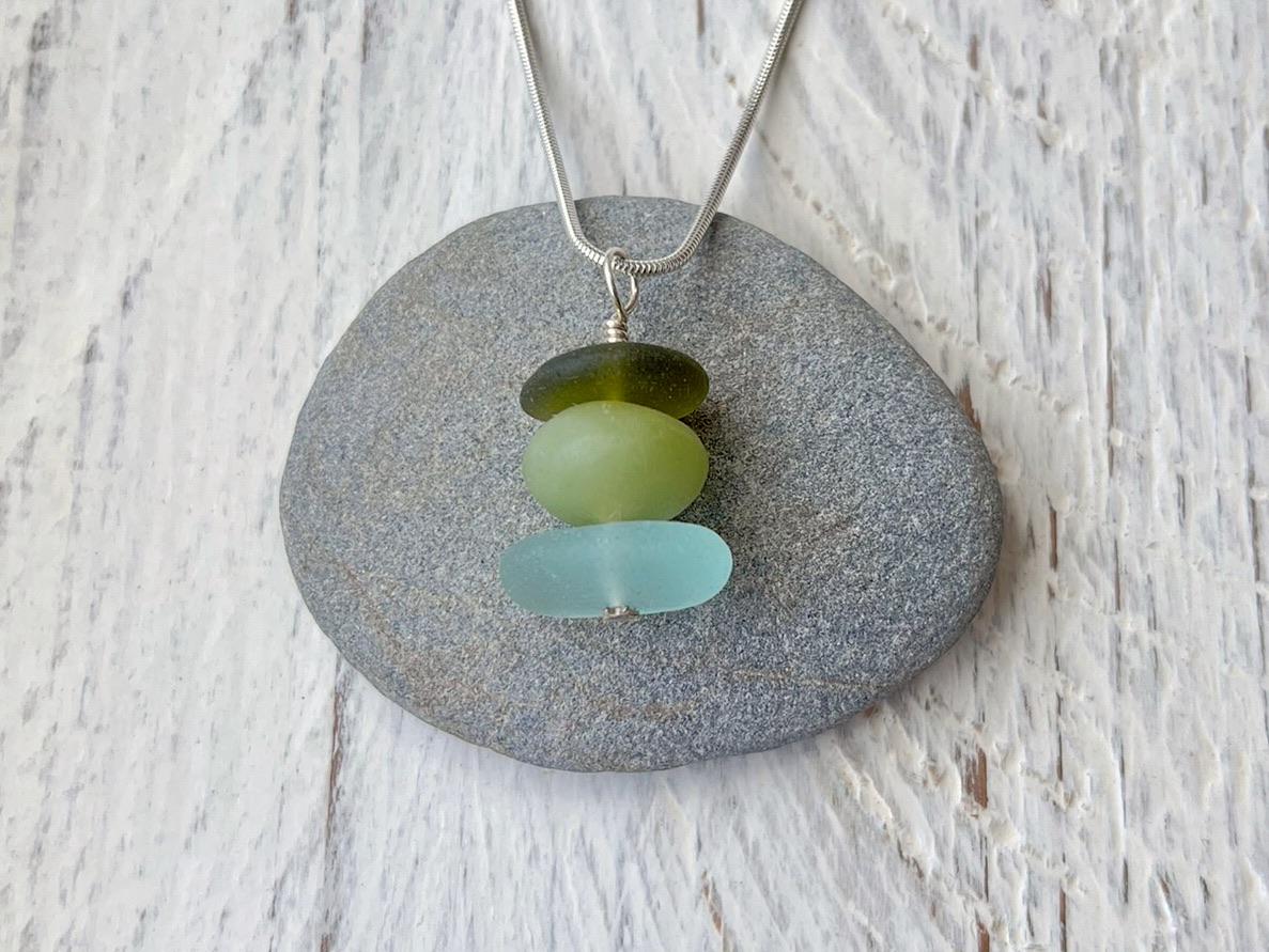 Necklace with green sea glass and a Jade bead laid on a grey pebble against a textured surface.