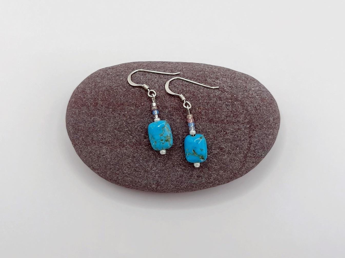 Turquoise beaded earrings laid on a purple pebble with a white background