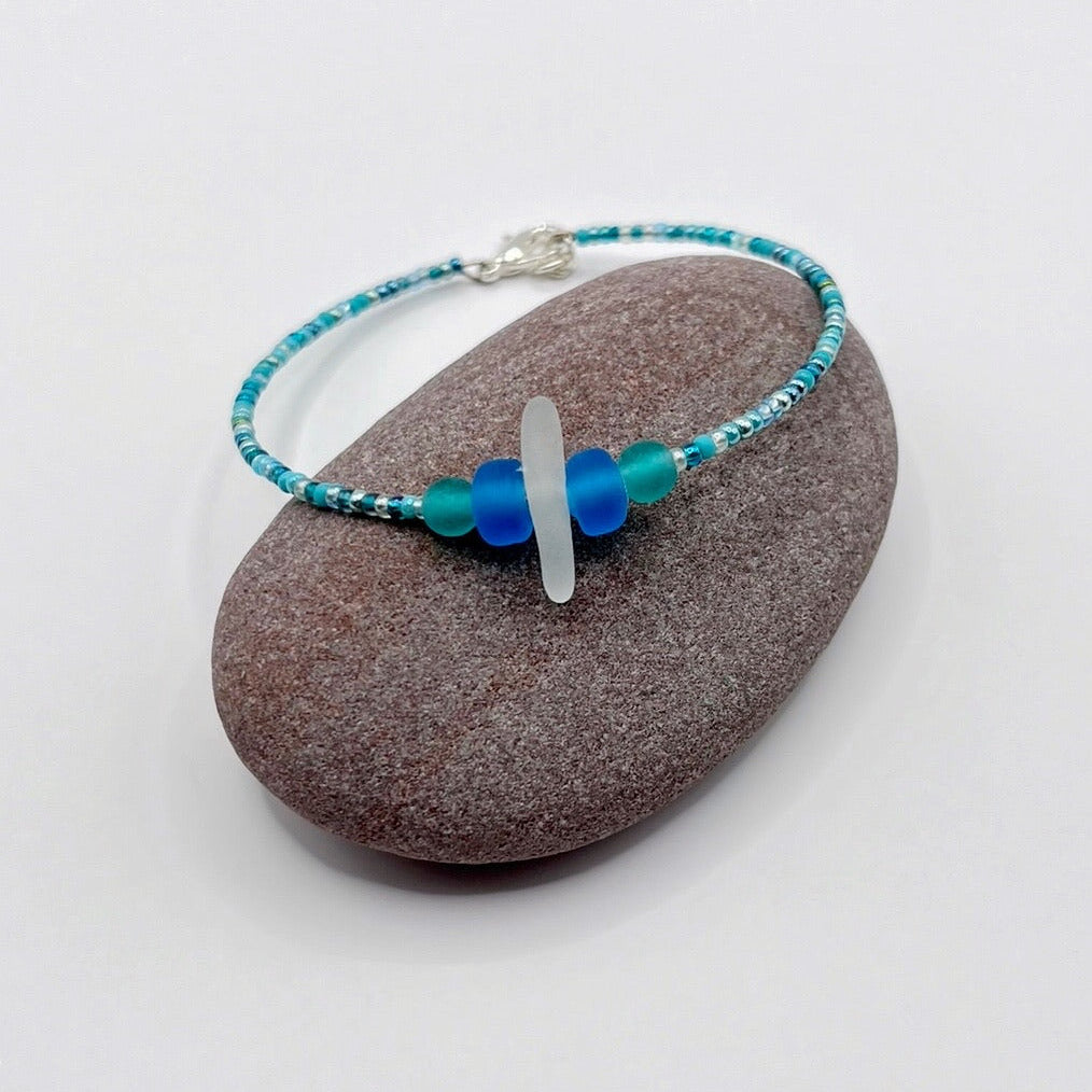 Bracelet with white sea glass and blue and turquoise beads laid on a purple pebble against a white background