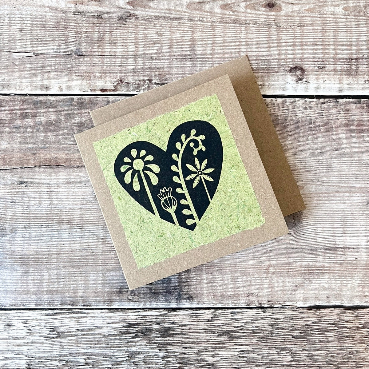 Square kraft greeting card with a black floral heart lino print on pale green handmade paper mounted on the front. Laid on a matching envelope on a wooden background.