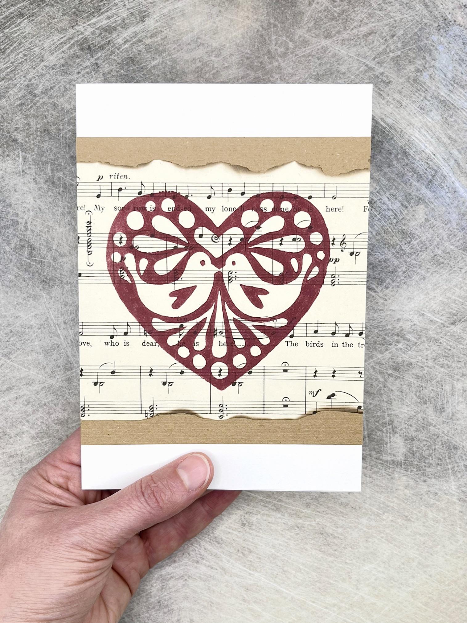 Card with original lino printed heart design on sheet music held by a hand against a textured grey background