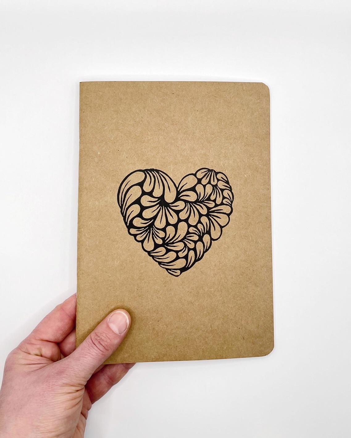 Kraft paper notebook with an original lino printed heart held by a hand against a white background