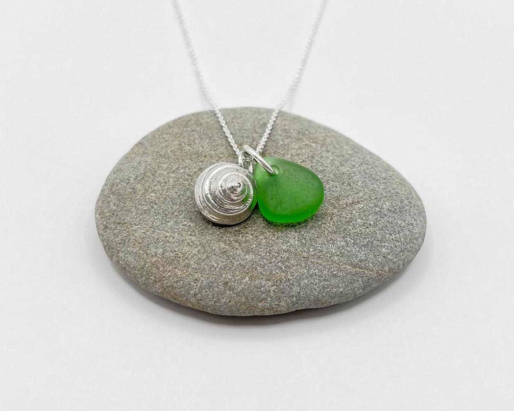Necklace with a silver shell and green sea glass laid on a grey pebble against a white background