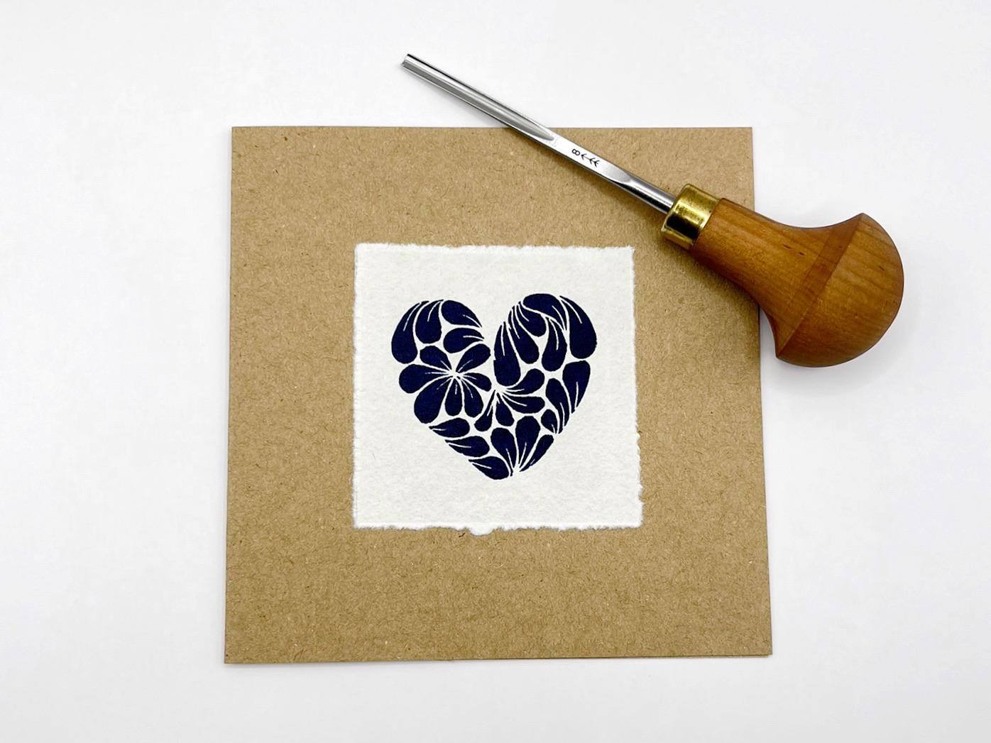 Kraft card with original lino printed blue heart design on white paper, laid on a white background. Photo includes lino cutting tool.