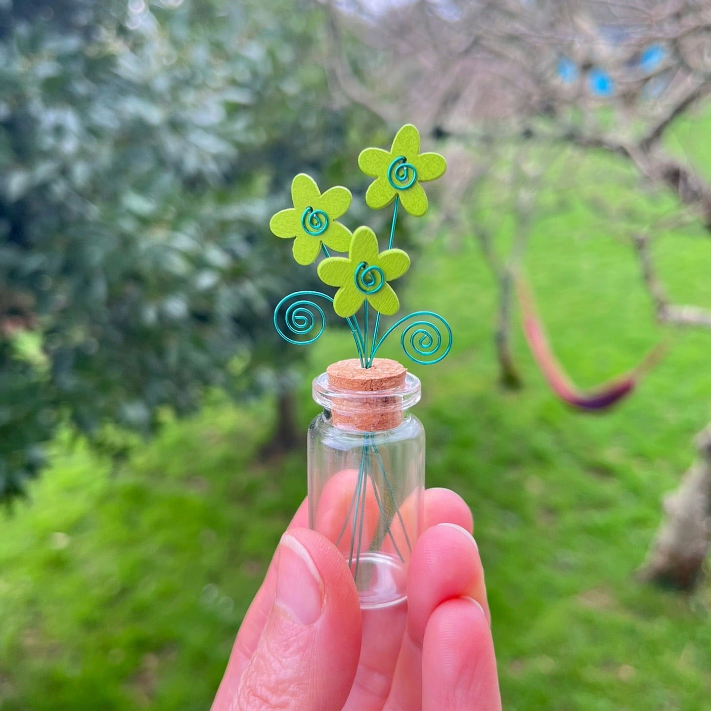Little glass bottle with three green wooden flowers held in a hand against a garden background