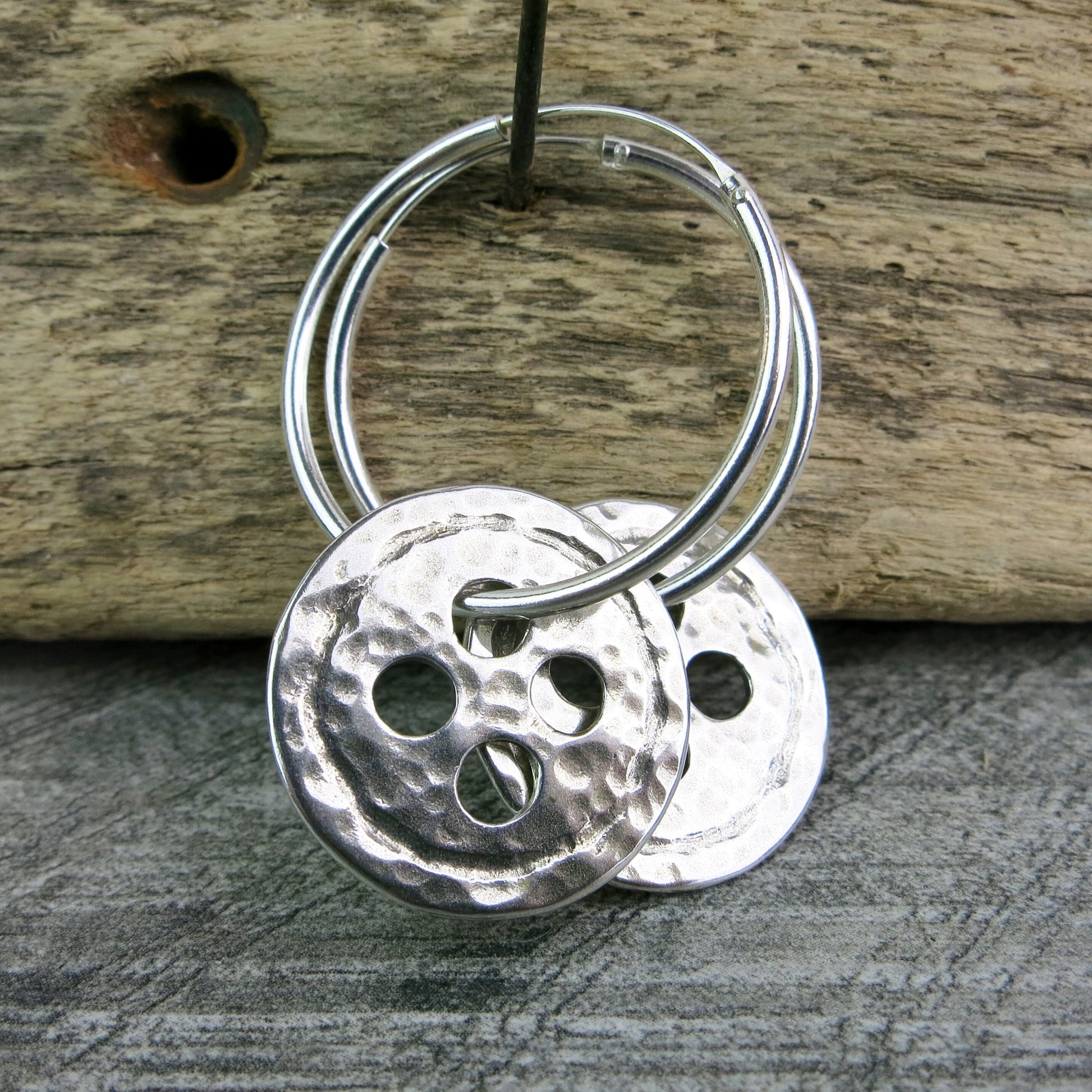 Silver hoop earrings with large round hammered silver buttons hanging on driftwood against a textured background