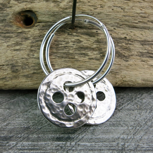 Silver hoop earrings with large round hammered silver buttons hanging on driftwood against a textured background