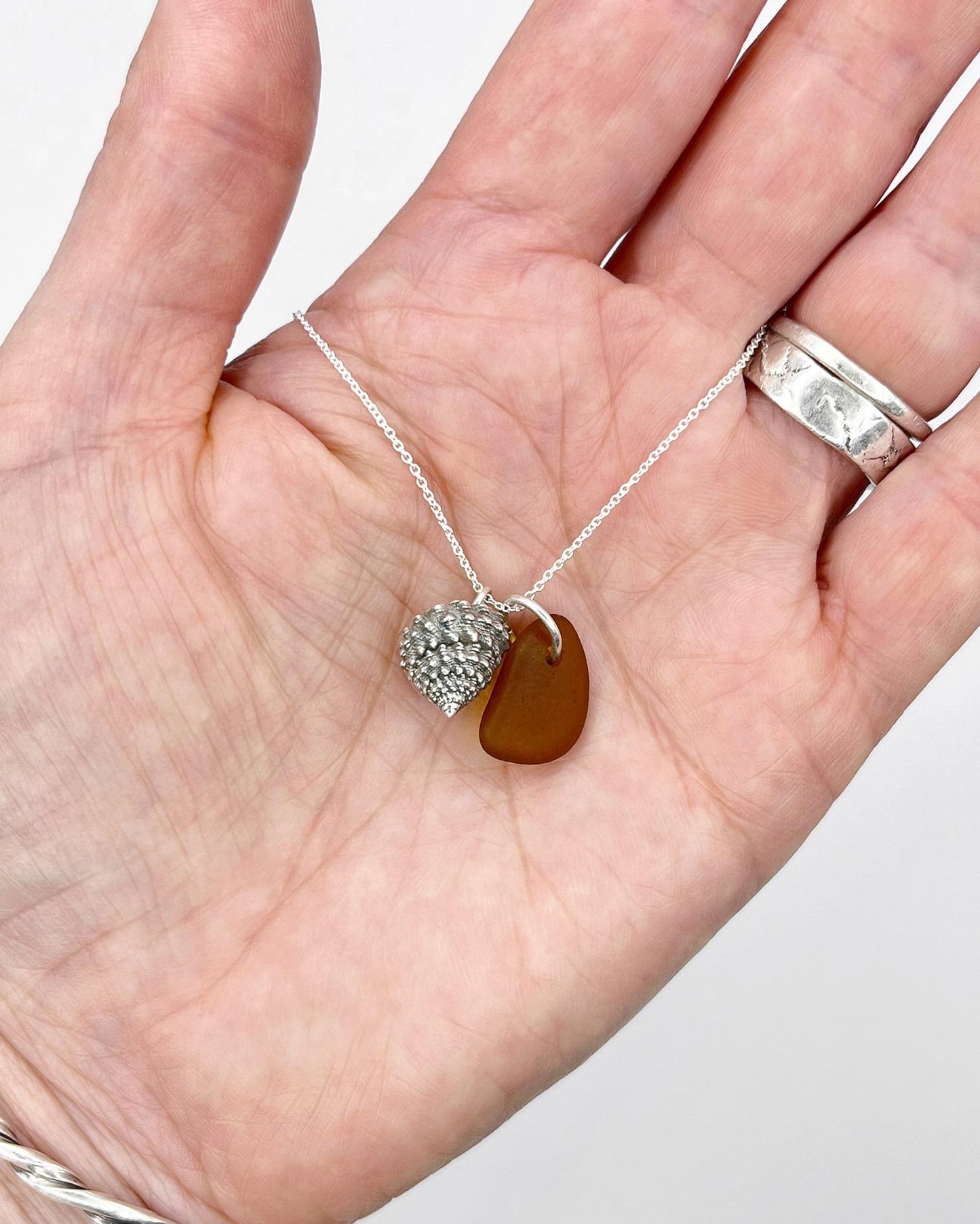 Necklace with burnt orange sea glass and silver shell pendant held in a hand against a white background