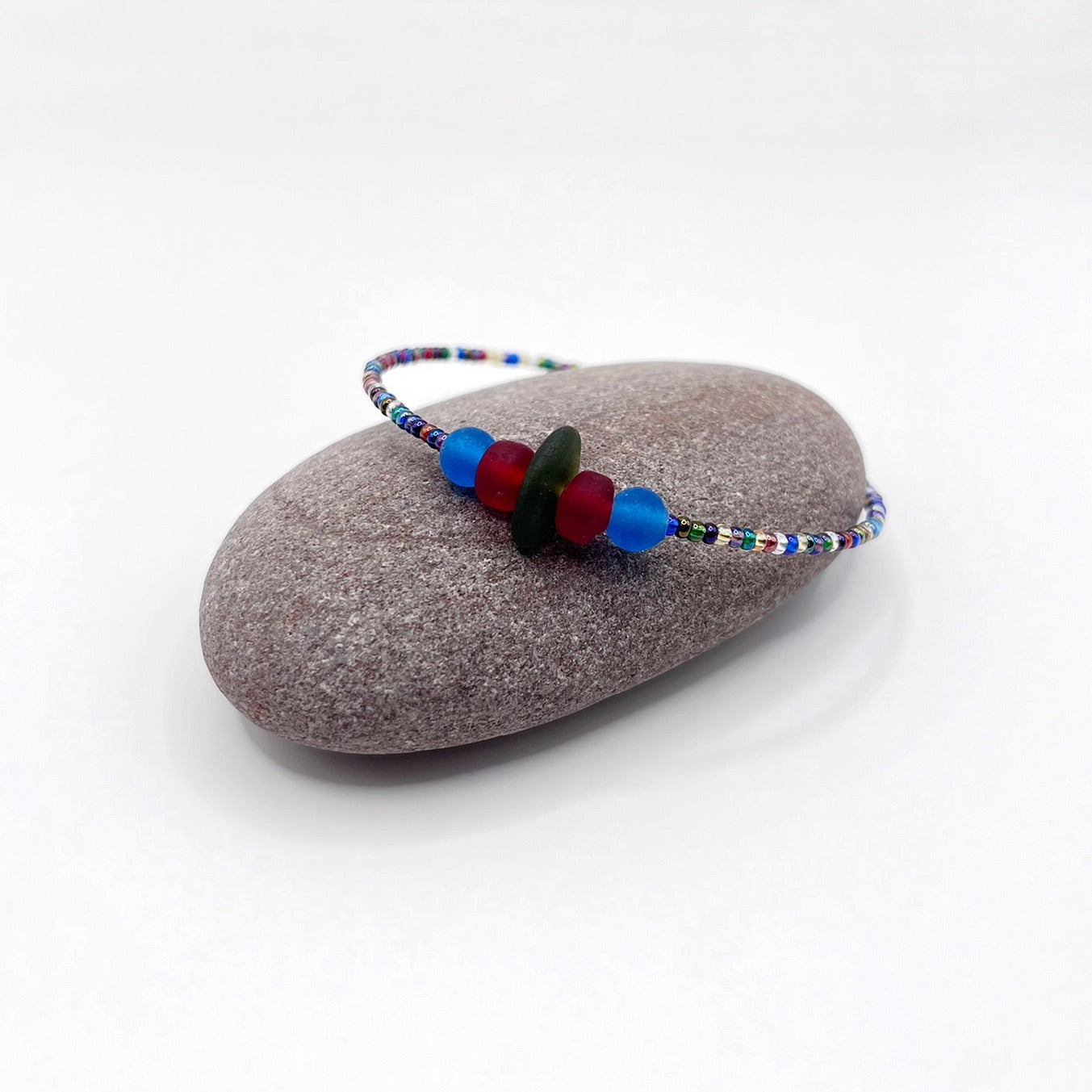 A rainbow coloured beaded bracelet with red and blue frosted glass beads and a piece of olive green coloured Cornish sea glass, finished with a silver plated lobster clasp and laid on a purple pebble.