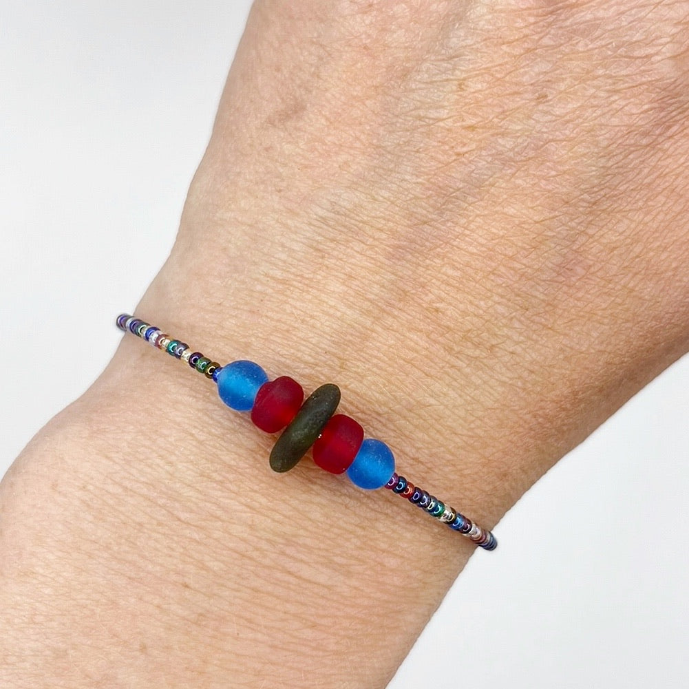 Close up of a rainbow coloured beaded bracelet with red and blue frosted glass beads and a piece of olive green coloured Cornish sea glass, worn on a wrist.