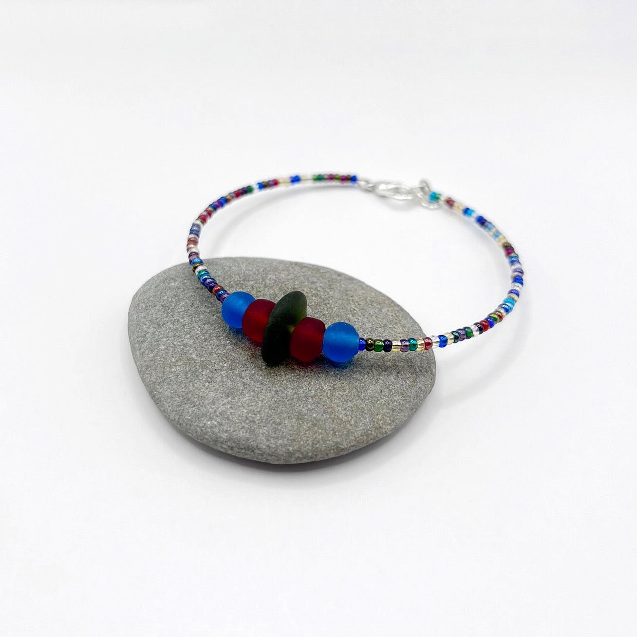A rainbow coloured beaded bracelet with red and blue frosted glass beads and a piece of olive green coloured Cornish sea glass, finished with a silver plated lobster clasp and laid on a grey pebble.