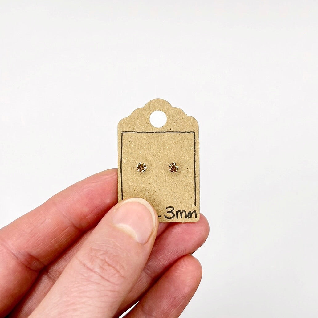 Small sparkly glass astud earrings on a cardboard display card held by a hand against a white background