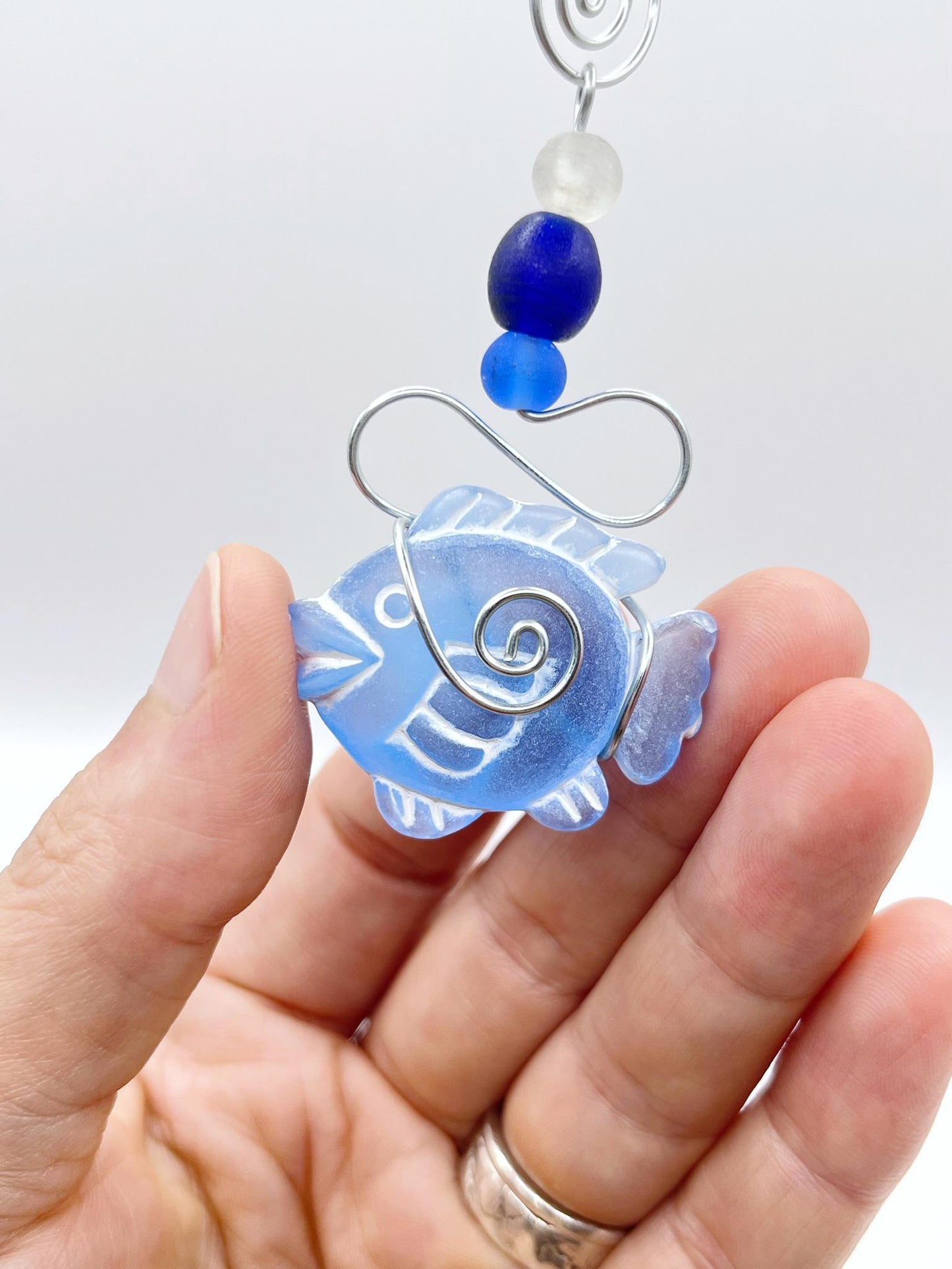 Close up of decorative hanging ornament with blue fish design, wire and glass beads held in a hand against a white background