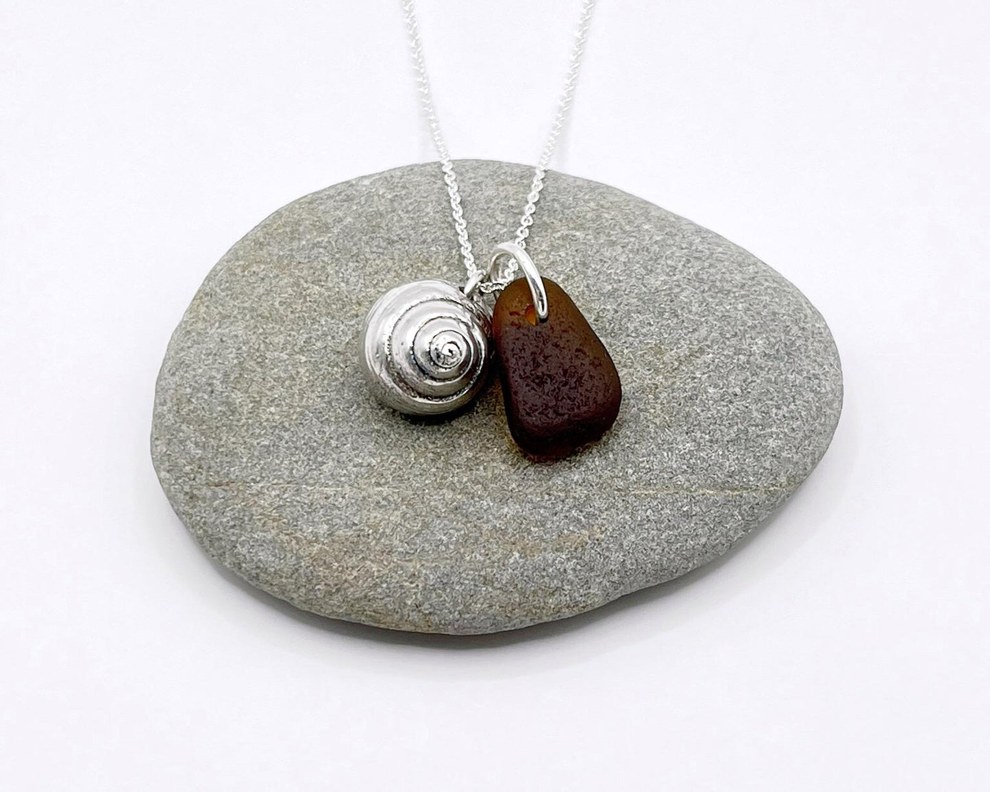 Silver necklace with burnt orange sea glass and a silver shell pendant laid on pebble against a white background