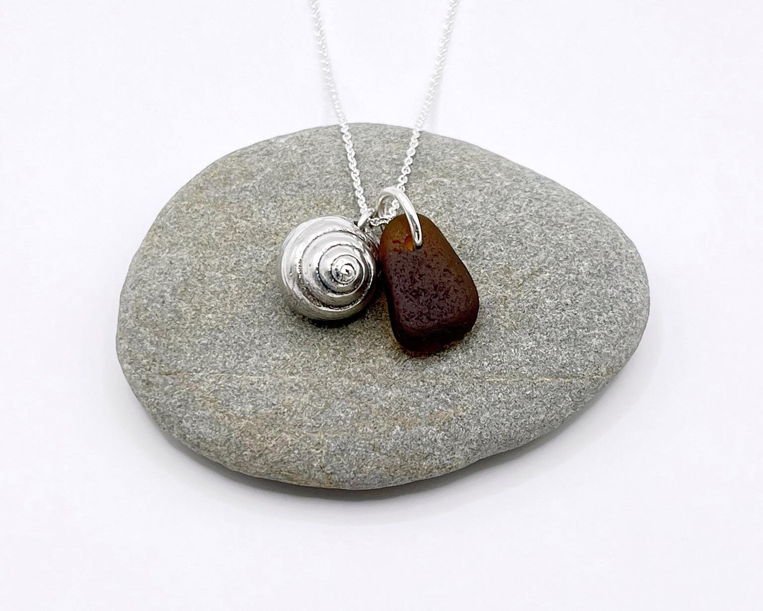 Silver necklace with burnt orange sea glass and a silver shell pendant laid on pebble against a white background