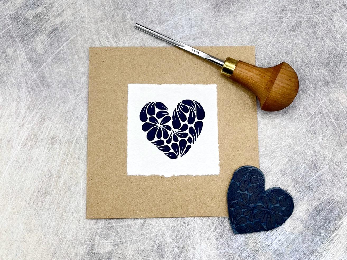 Kraft card with original lino printed blue heart design on white paper, laid on a textured grey background. Photo includes lino and cutting tool.