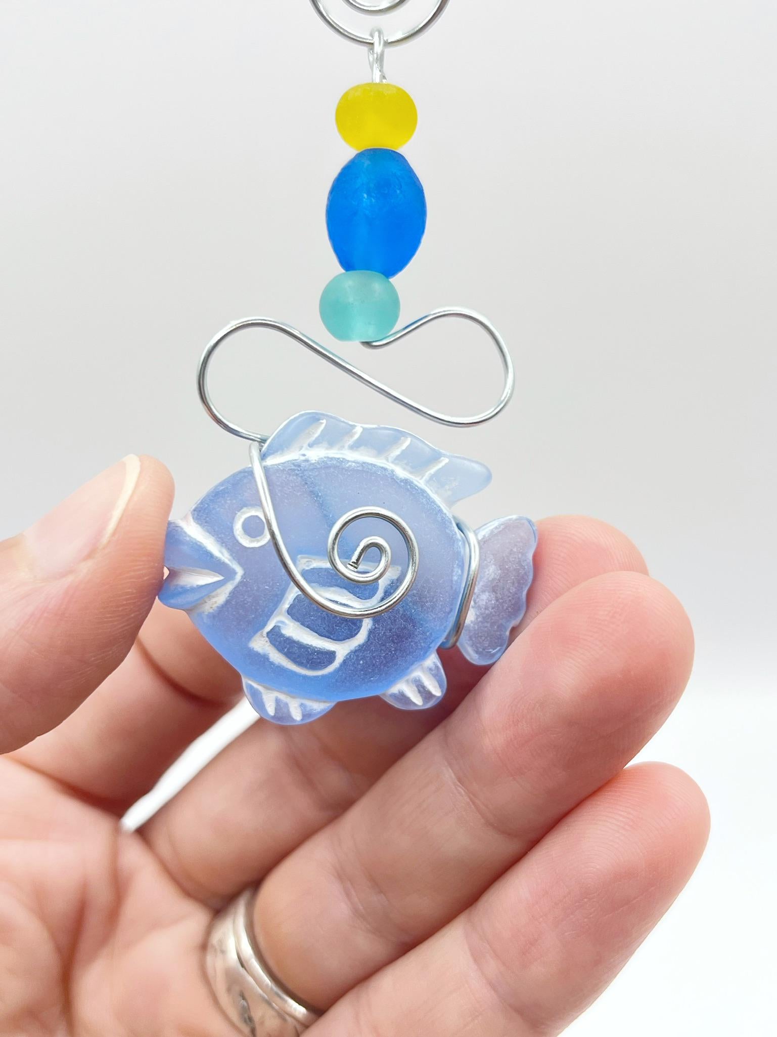Close up of decorative ornament with blue fish design, wire and colourful beads held in a hand against a white background