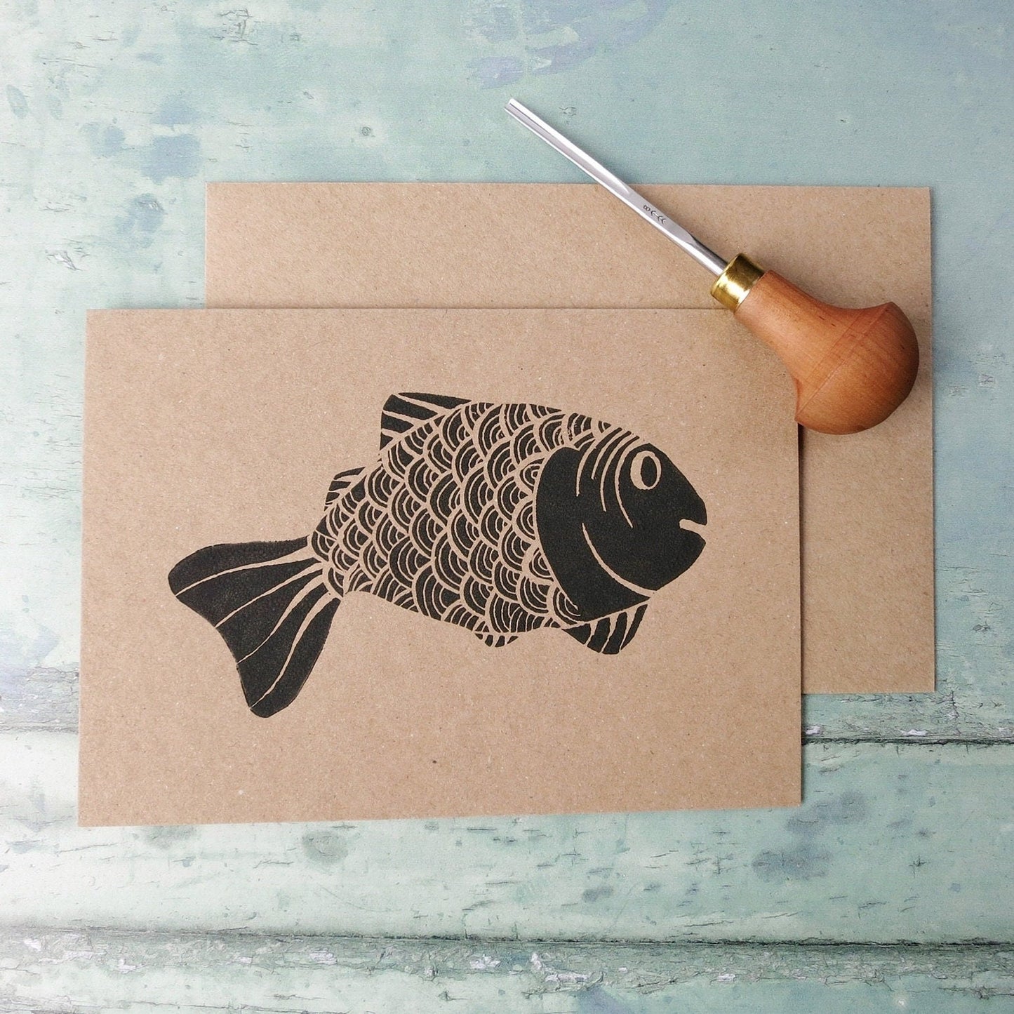Kraft paper card with original lino printed fish laid on a textured surface. Photo includes lino cutting tool