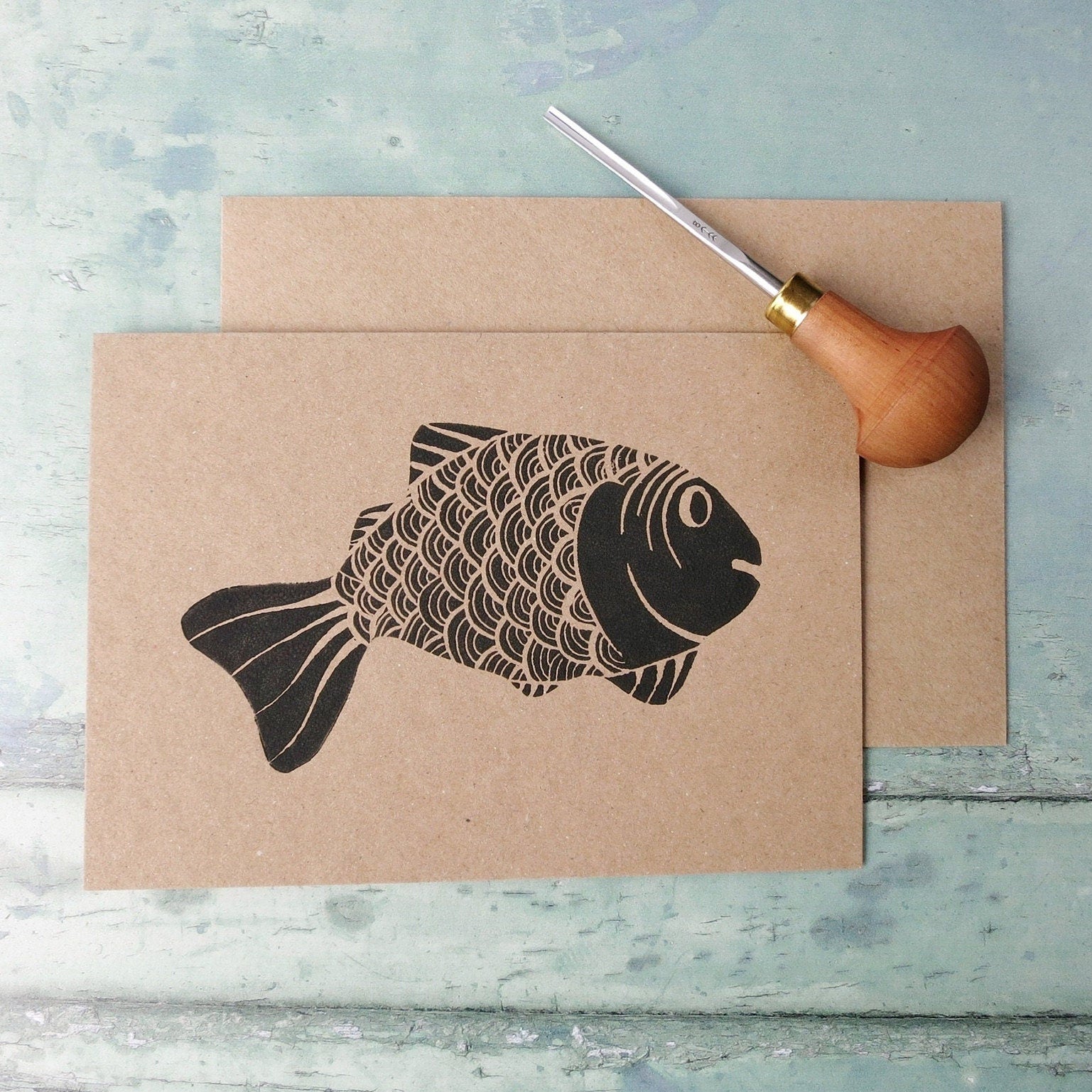 Kraft paper card with original lino printed fish laid on a textured surface. Photo includes lino cutting tool
