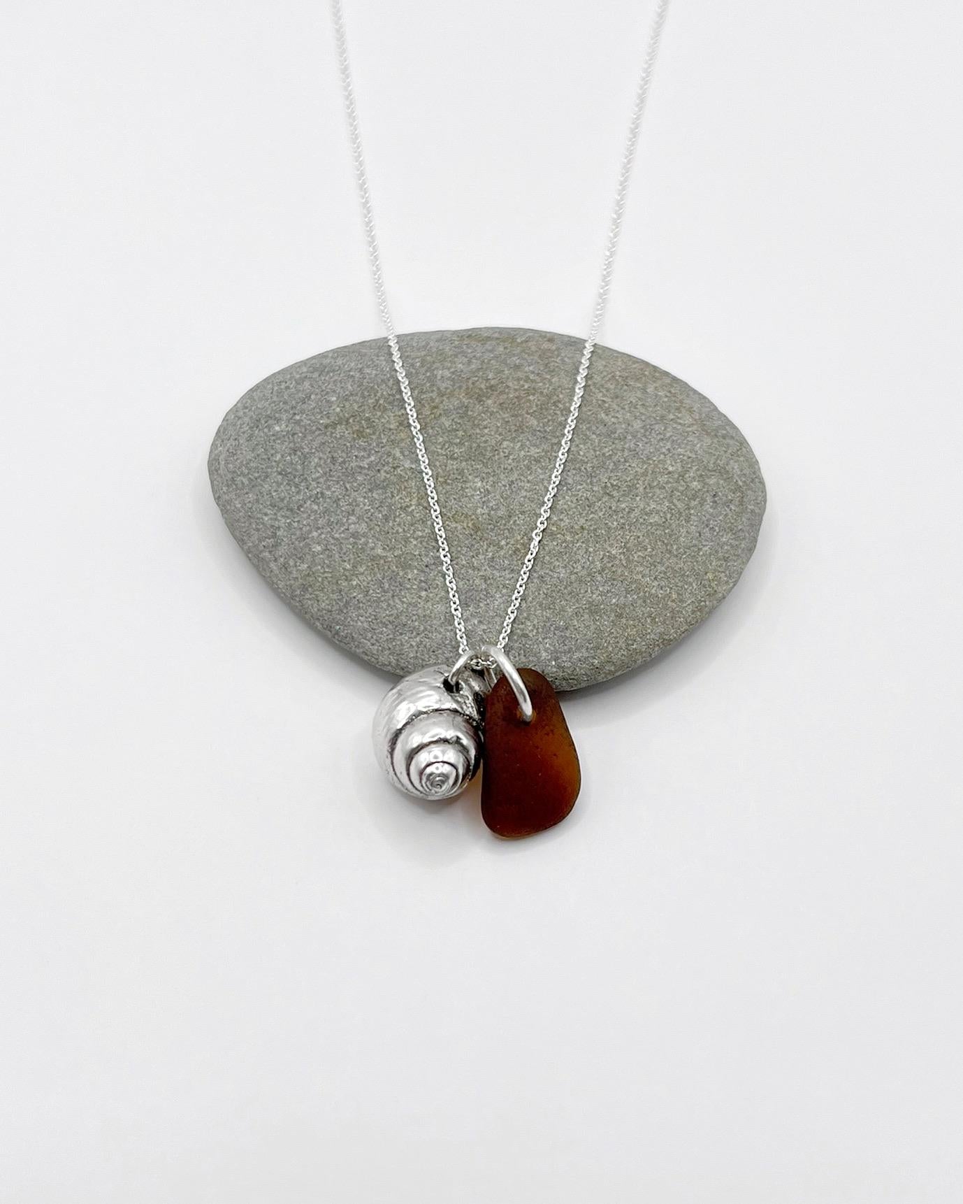 Silver necklace with burnt orange sea glass and a silver shell pendant laid on pebble against a white background