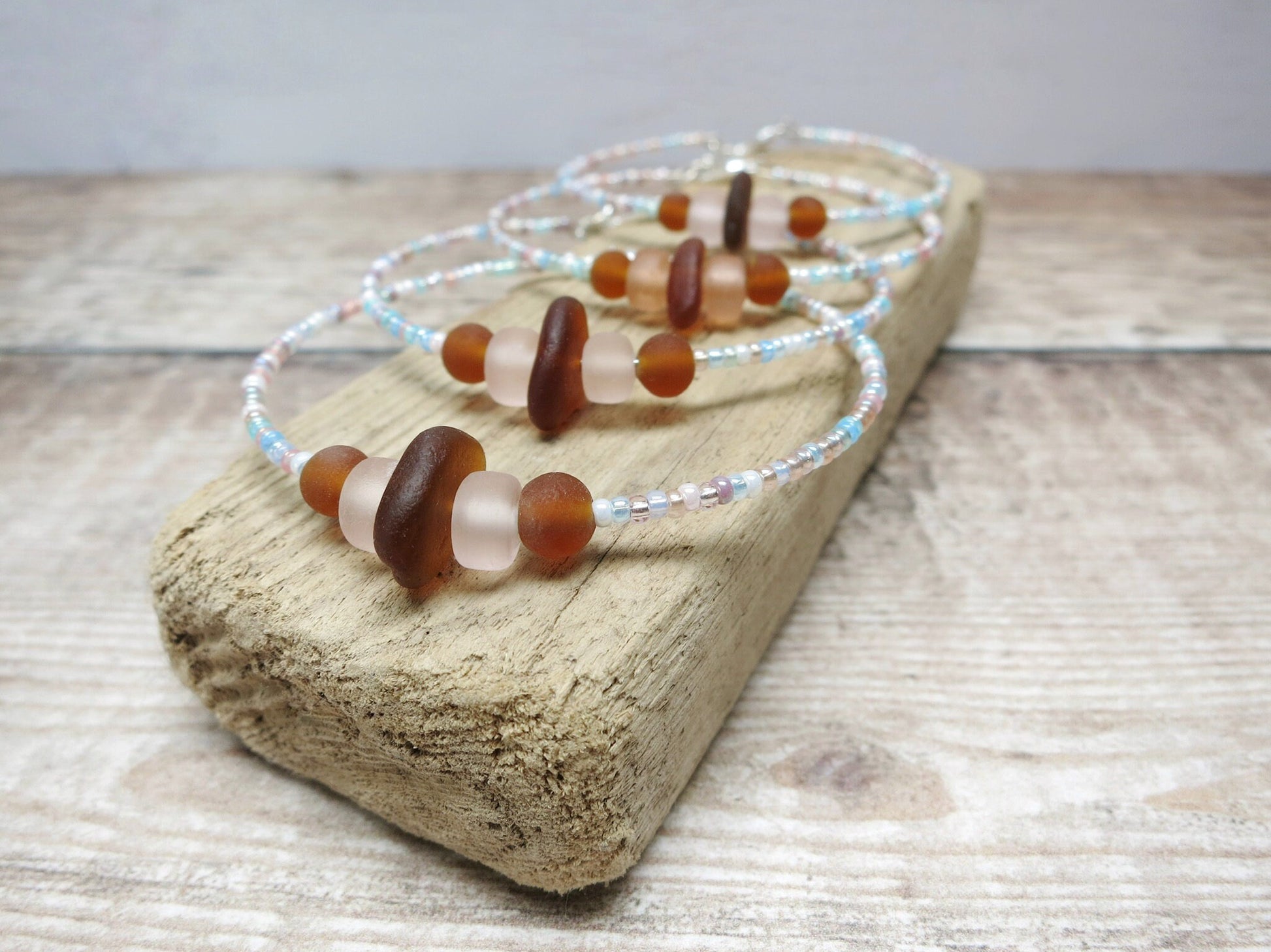 Close up of bracelets with pastel seed beads and burnt orange sea glass laid on driftwood.