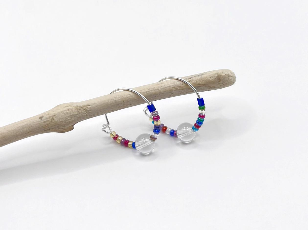 Rainbow seed bead and crystal hoop earrings hanging on driftwood against a white background.