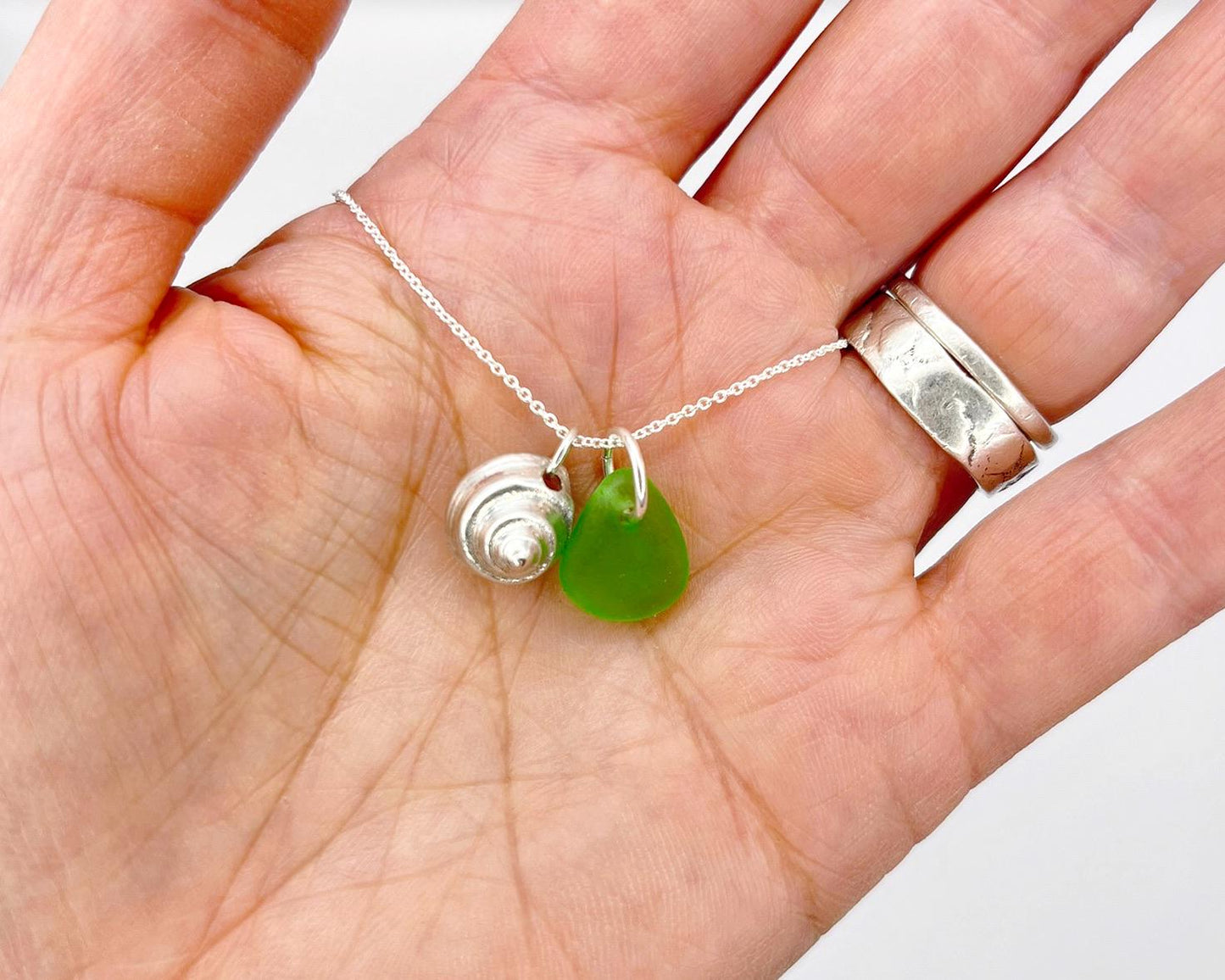Hand holding a necklace with a green sea glass and silver shell pendant against a white background