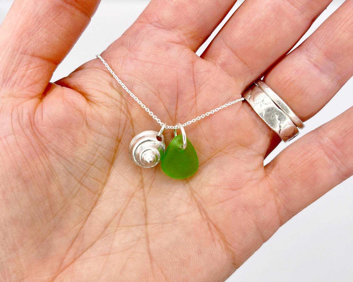 Hand holding a necklace with a green sea glass and silver shell pendant against a white background